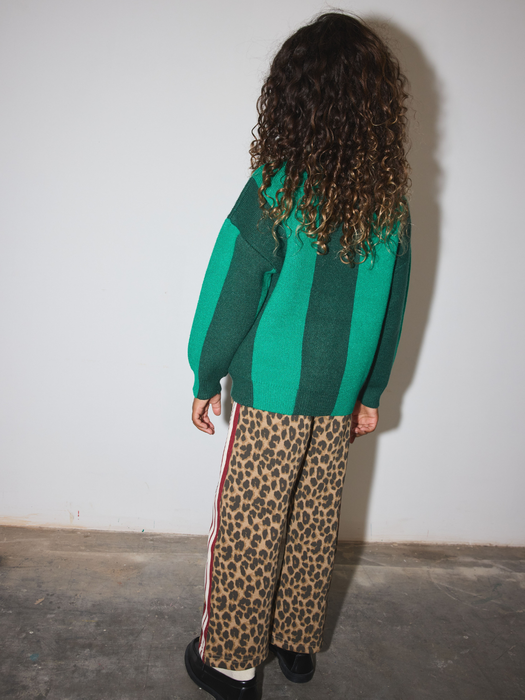 A child standing with their back to the camera, wearing a green and dark green striped knit sweater and leopard print pants with a red side stripe.