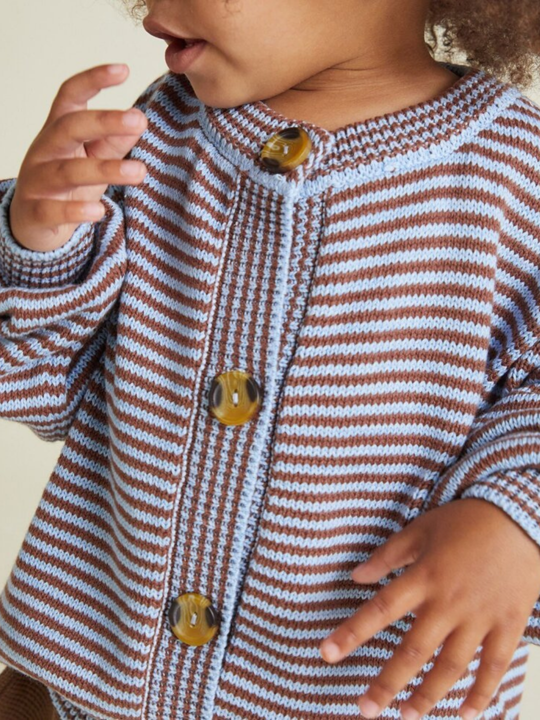 A young child wears the EMIL COCO SKY STRIPE CARDIGAN—a brown and light blue striped knit sweater with large yellow buttons; only the lower part of the child's face and upper body are shown.