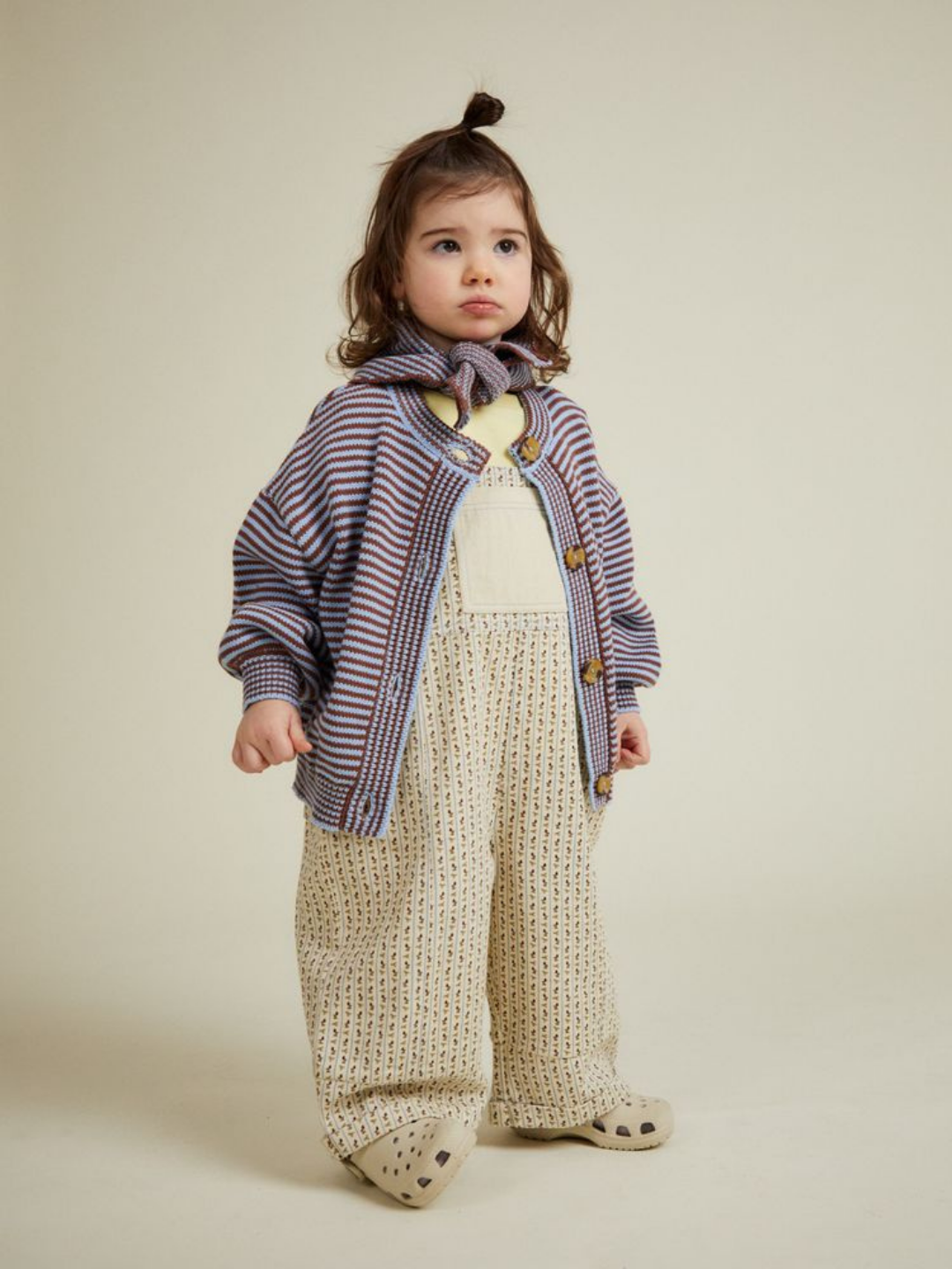 A young child stands facing forward against a plain background, wearing the EMIL COCO SKY STRIPE CARDIGAN with loose patterned overalls, a scarf, and light-colored shoes.