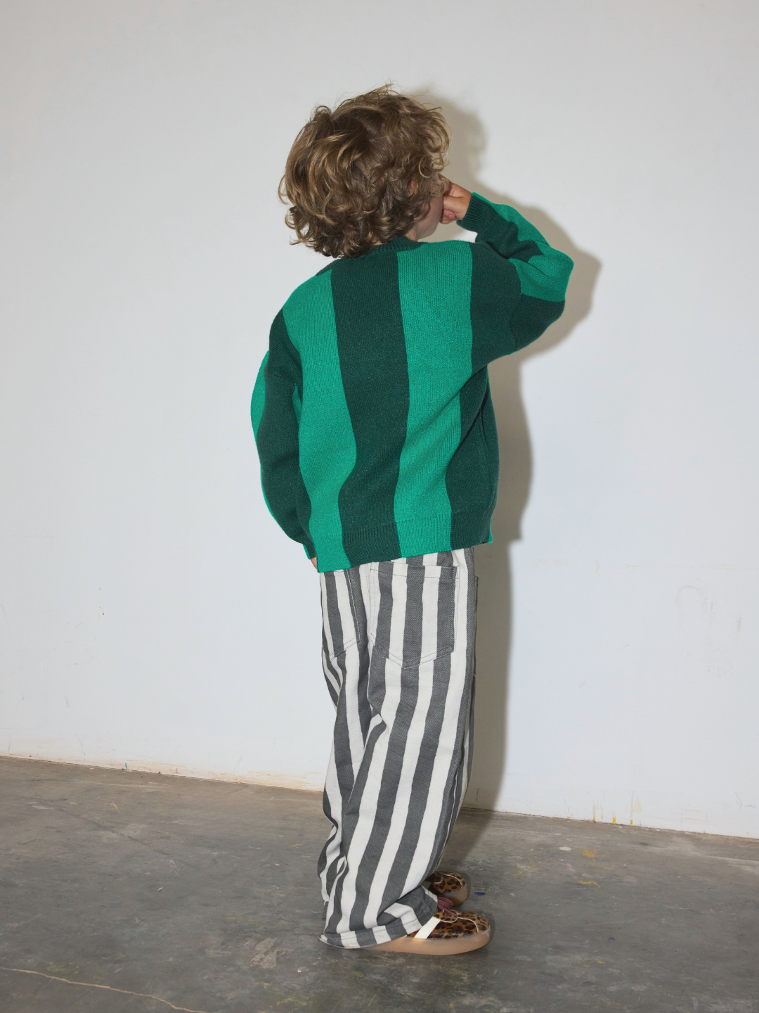 Green | A child standing with their back to the camera, wearing a green and dark green striped knit cardigan and black and white vertically striped pants.