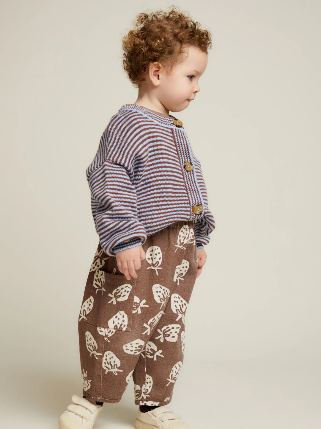 Toddler standing sideways in the EMIL COCO SKY STRIPE CARDIGAN, paired with brown pants featuring a white tree pattern and cream shoes, against a plain background.