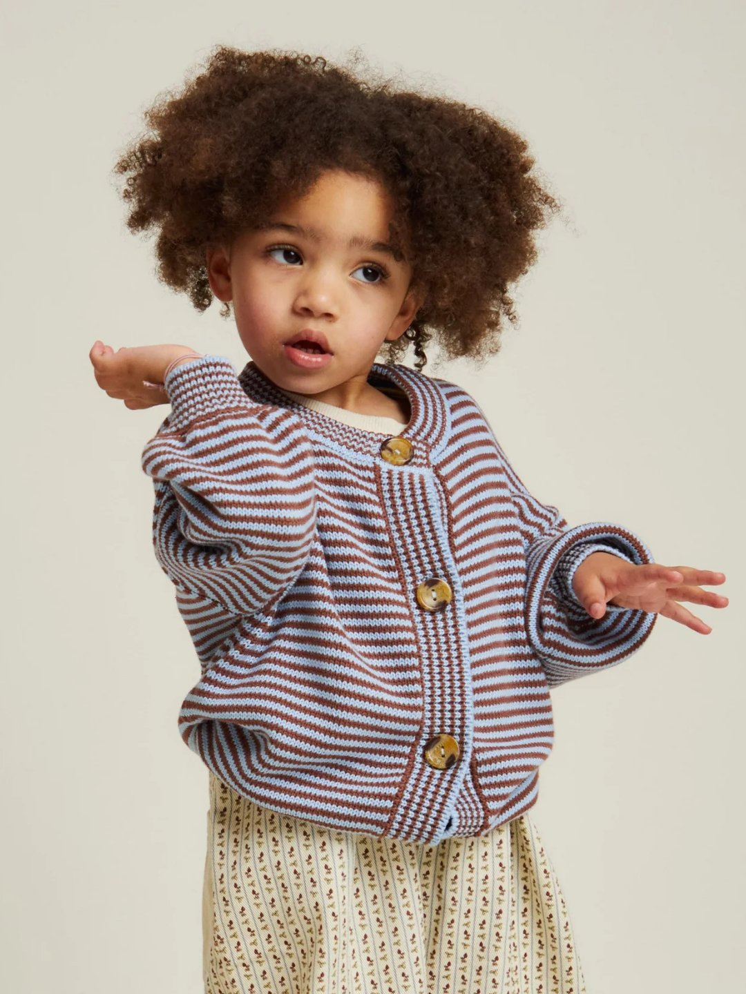 Young child with curly hair wearing the EMIL COCO SKY STRIPE CARDIGAN, standing against a plain background with one arm raised and the other extended.