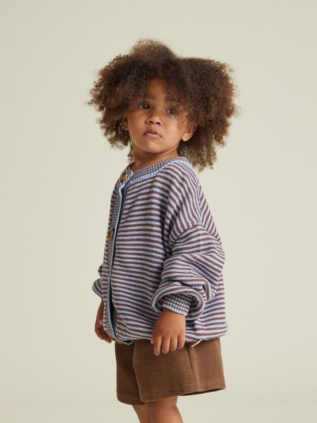 A young child with curly hair wears the EMIL COCO SKY STRIPE CARDIGAN and brown shorts, standing against a plain light background.