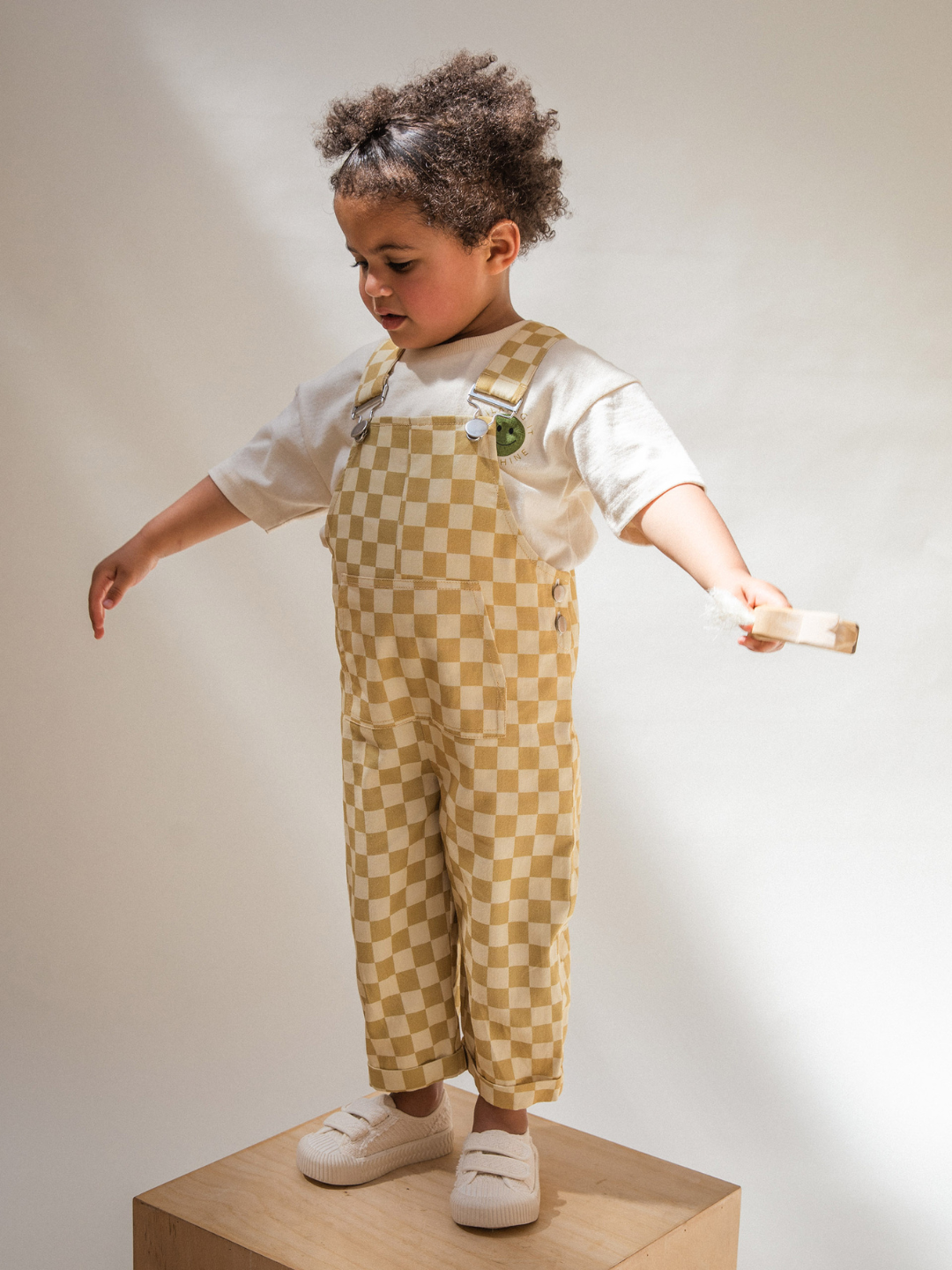 A young child with curly hair stands on a wooden box, wearing the Check Ochre Dungaree in 100% organic cotton by Claude & Co over a white shirt, holding a light-colored wooden toy. The child appears to be balancing with arms outstretched against a plain white background, creating soft, natural light.