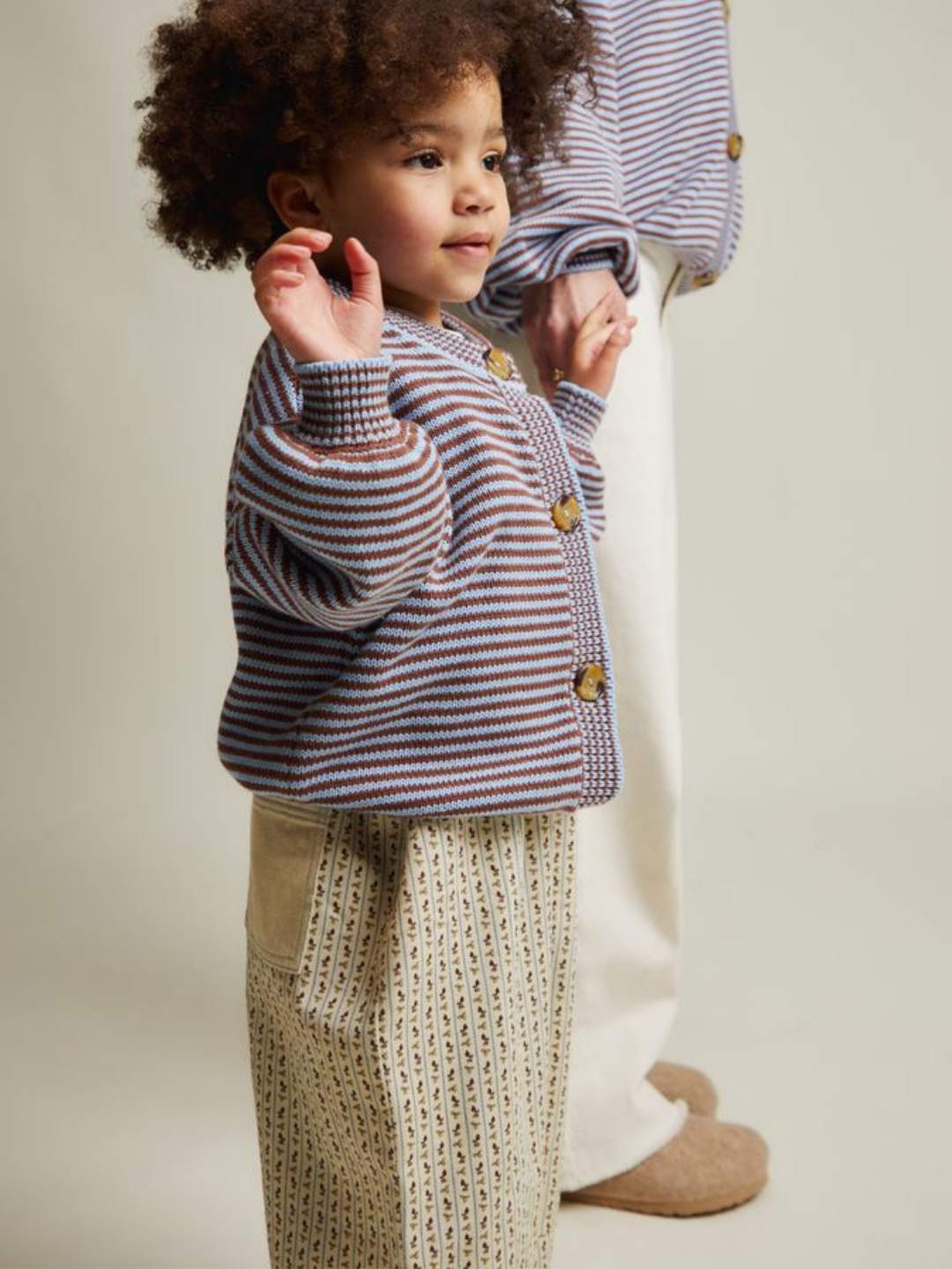 Young child in profile wears the EMIL COCO SKY STRIPE CARDIGAN with patterned pants, beside a partially visible adult, in a neutral studio setting.