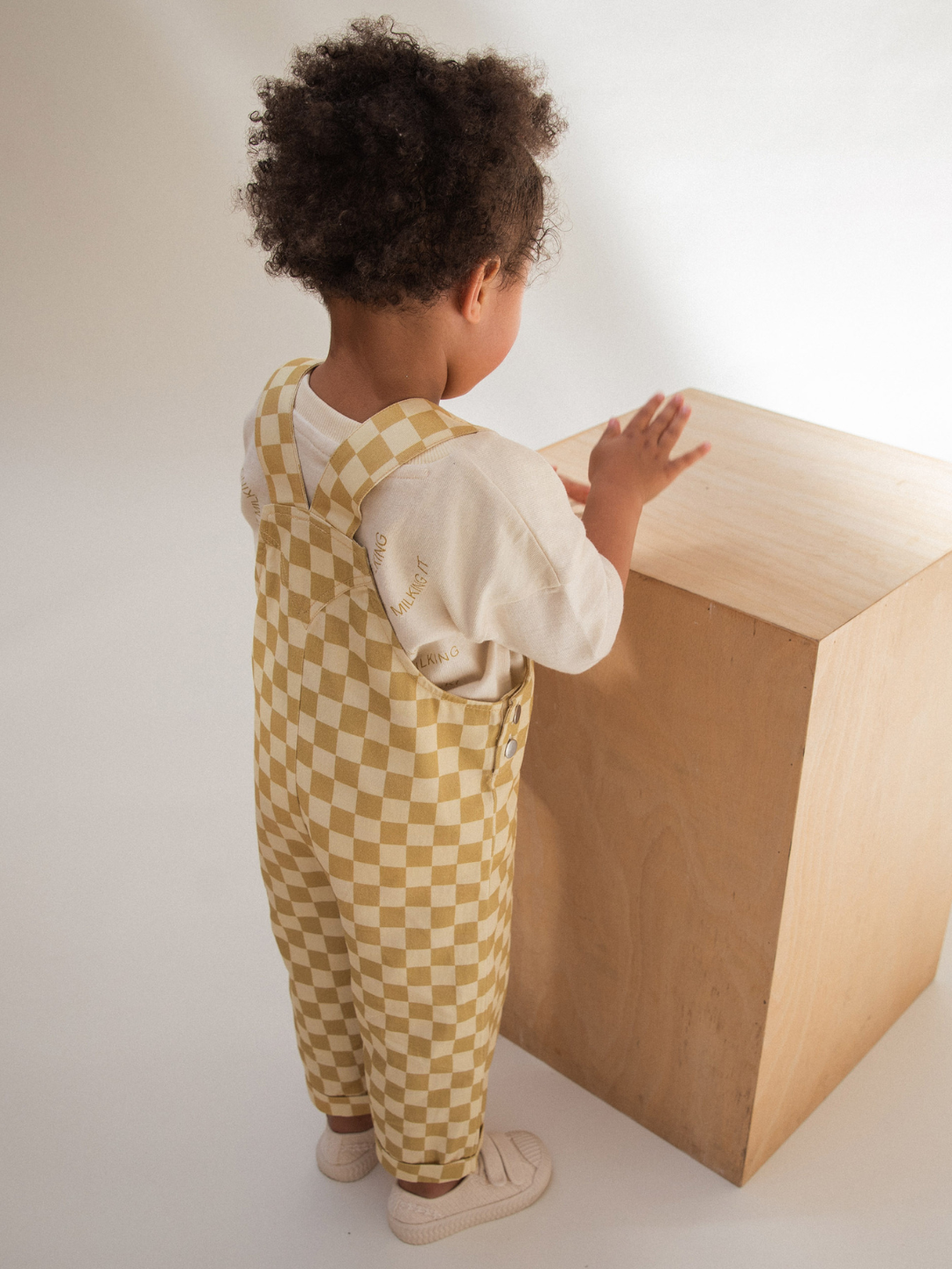 A young child with curly hair stands facing a wooden cube, wearing a white shirt and Claude & Co's CHECK OCHRE DUNGAREE made from 100% organic cotton. They are touching the cube with one hand. The background is plain and well-lit, highlighting the child and the cube.