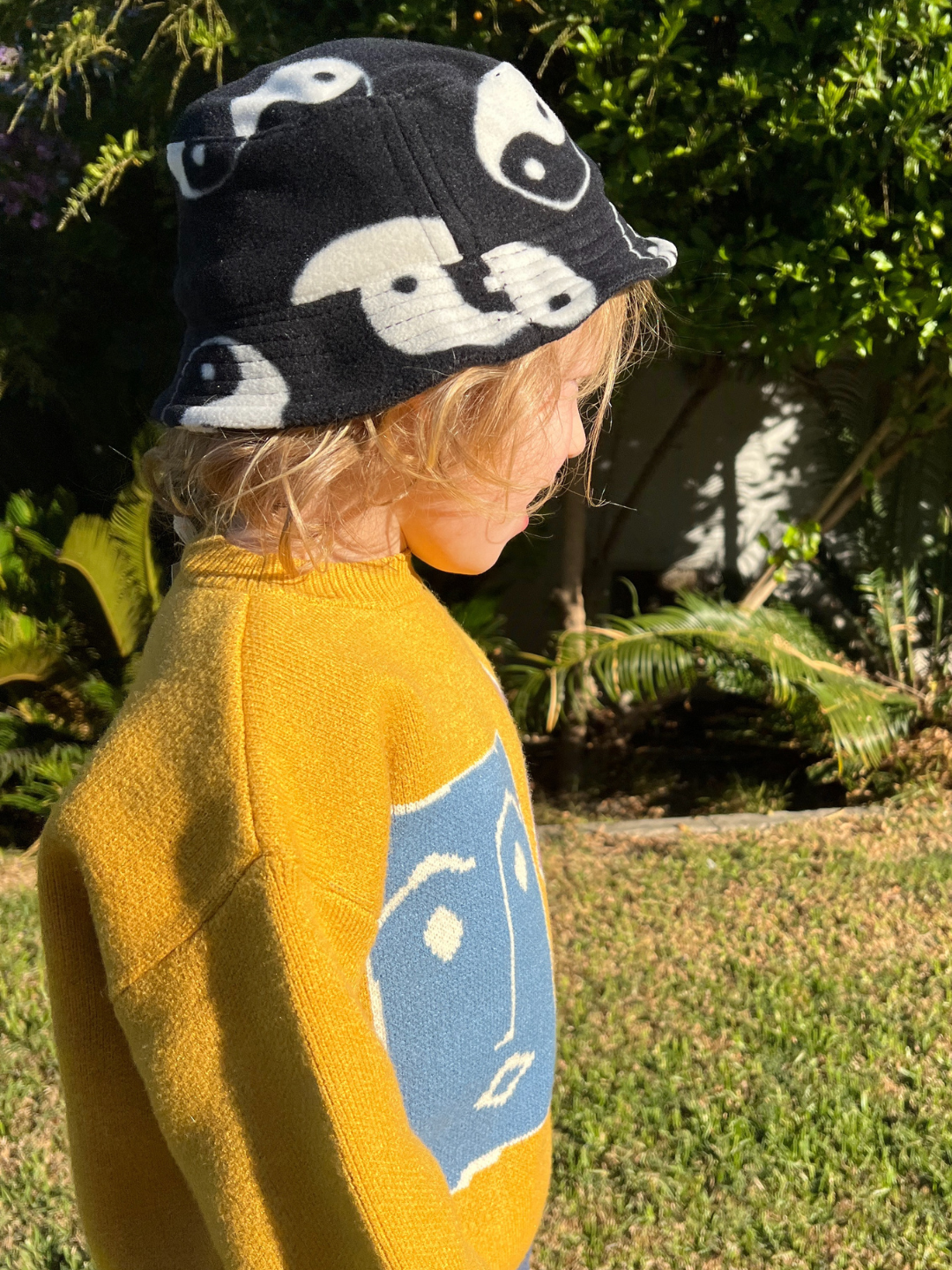 A child wearing the yin yang fleece bucket hat in black patterned all over with white yin yangs. They wear a yellow sweater with a blue square face design, and are standing in a green garden.