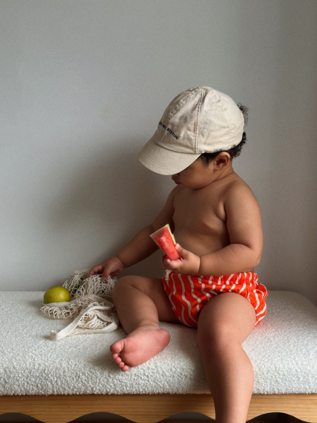 Spaghetti | The front view of the swim diaper with an elastic waist with a tie and elastic leg holes on a child sitting down. The diaper is a bright orange with wavy and vertical cream colored lines.