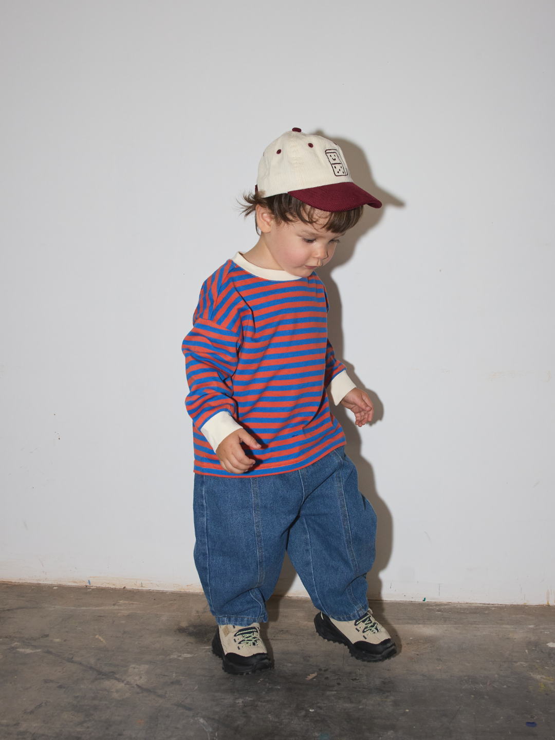 Red/Blue | A toddler standing against a plain off-white wall, wearing a cream baseball cap with a burgundy brim, a red and blue horizontally striped long-sleeve shirt, loose blue jeans, and beige-and-black sneakers.