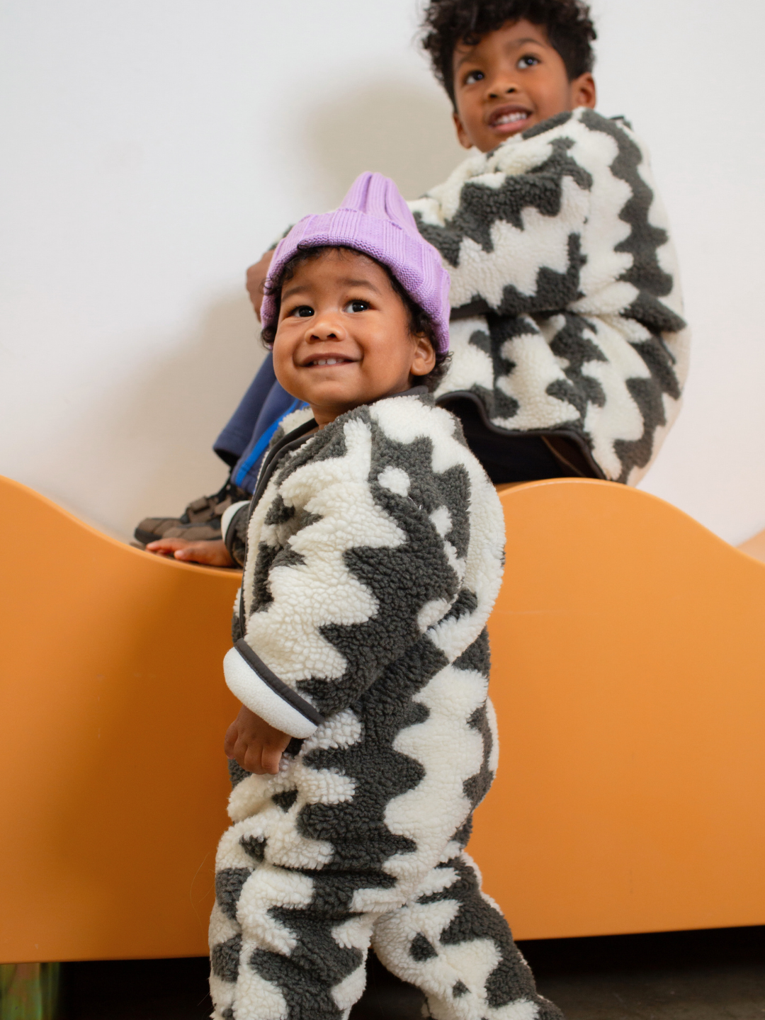 Charcoal | Two children wearing patterned outerwear sitting on a yellow surface.