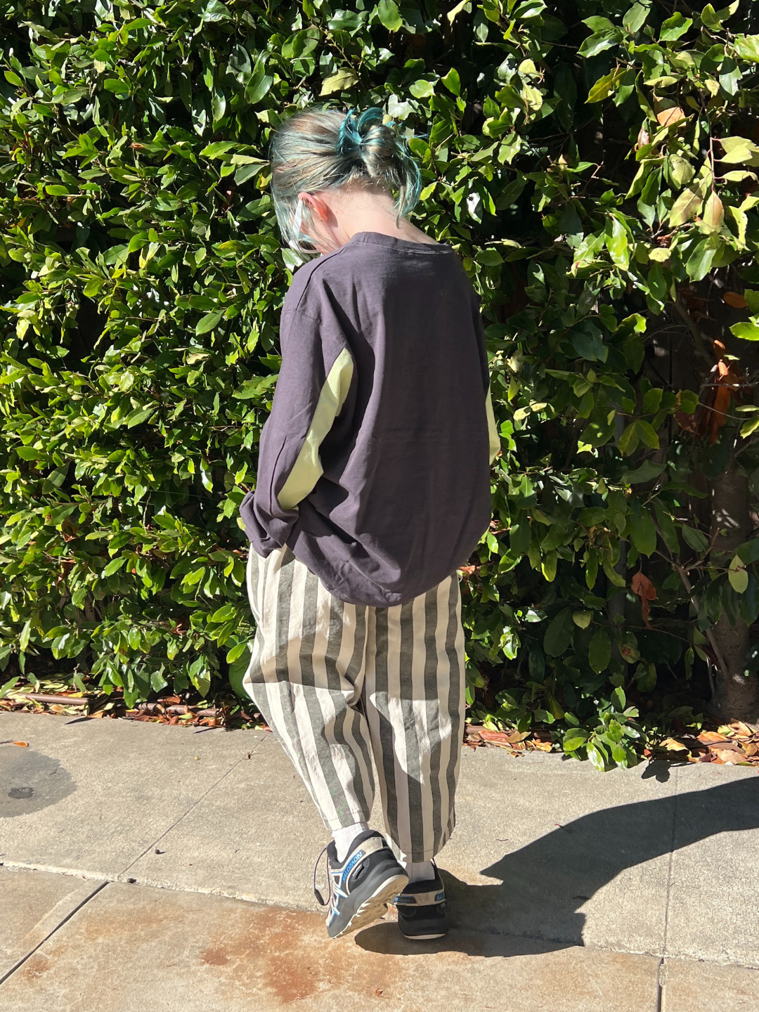 Person with striped pants and a dark shirt walking on a sidewalk with greenery in the background