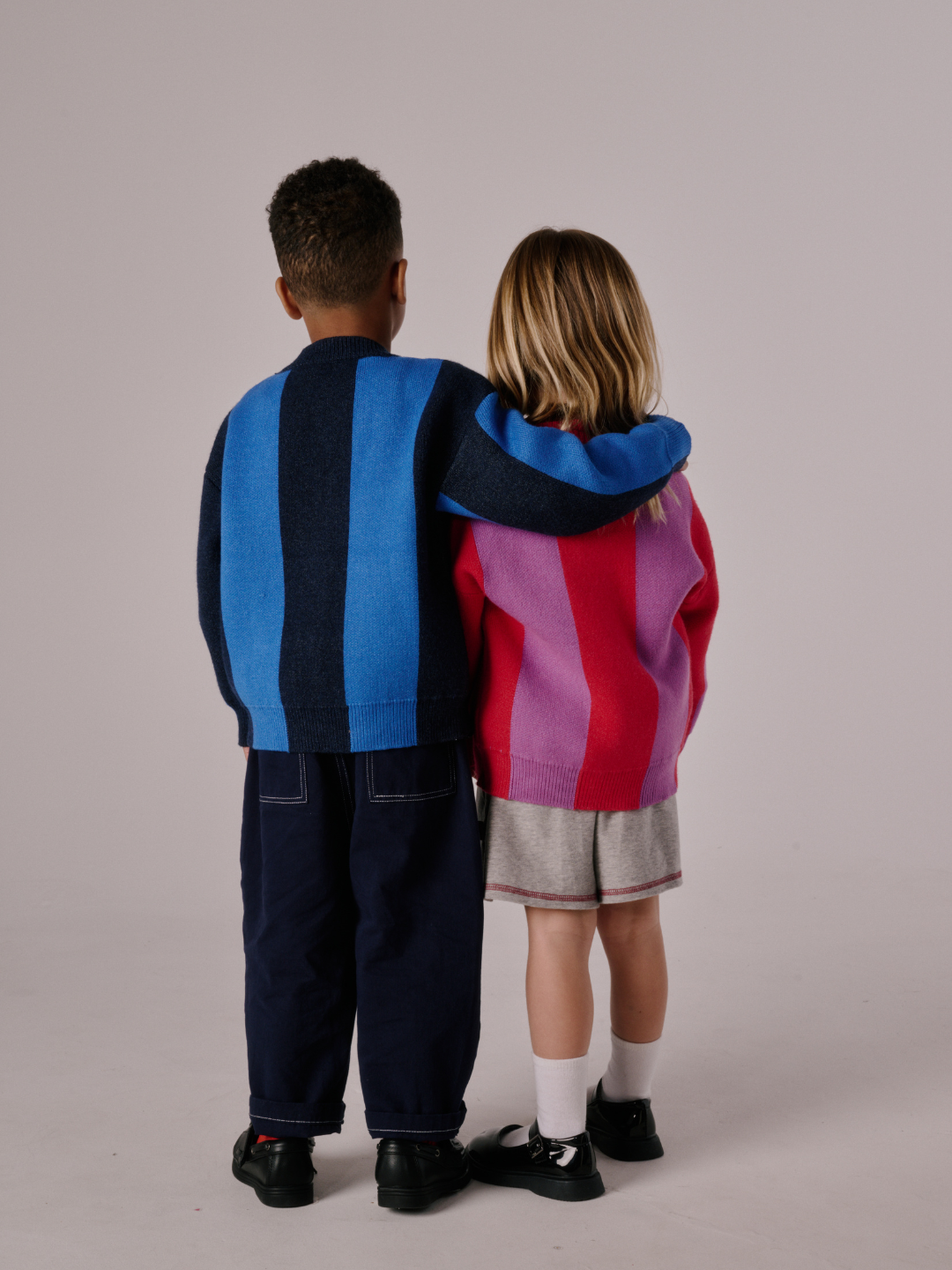 Two children stand side by side with their backs to the camera, one with an arm around the other's shoulders, both wearing STRIPE CARDIGAN sweaters.