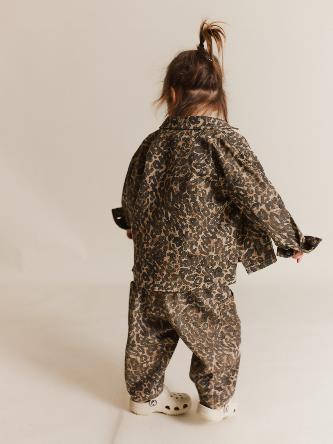 A toddler, wearing a FEN LEOPARD JACKET that's oversized and a pair of white Crocs, stands with their back to the camera.