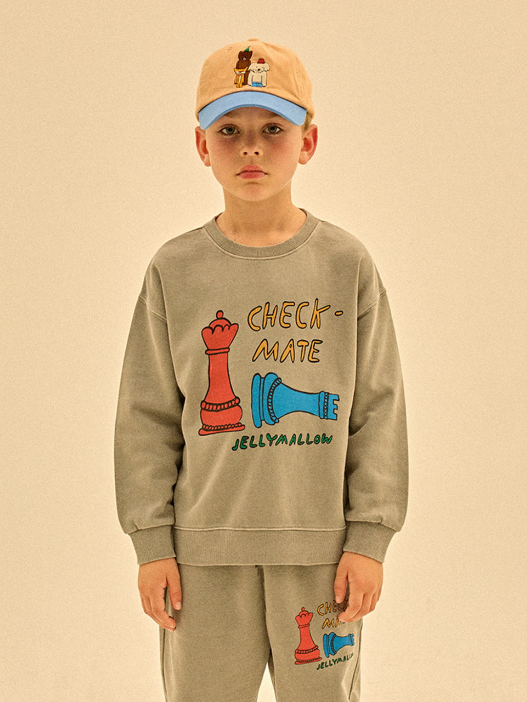 A young boy wears the CHECKMATE PIGMENT SWEATSHIRT in grey, featuring colorful chess piece graphics and "CHECKMATE JELLYMALLOW"—a playful look that stands out, with no added tariffs.