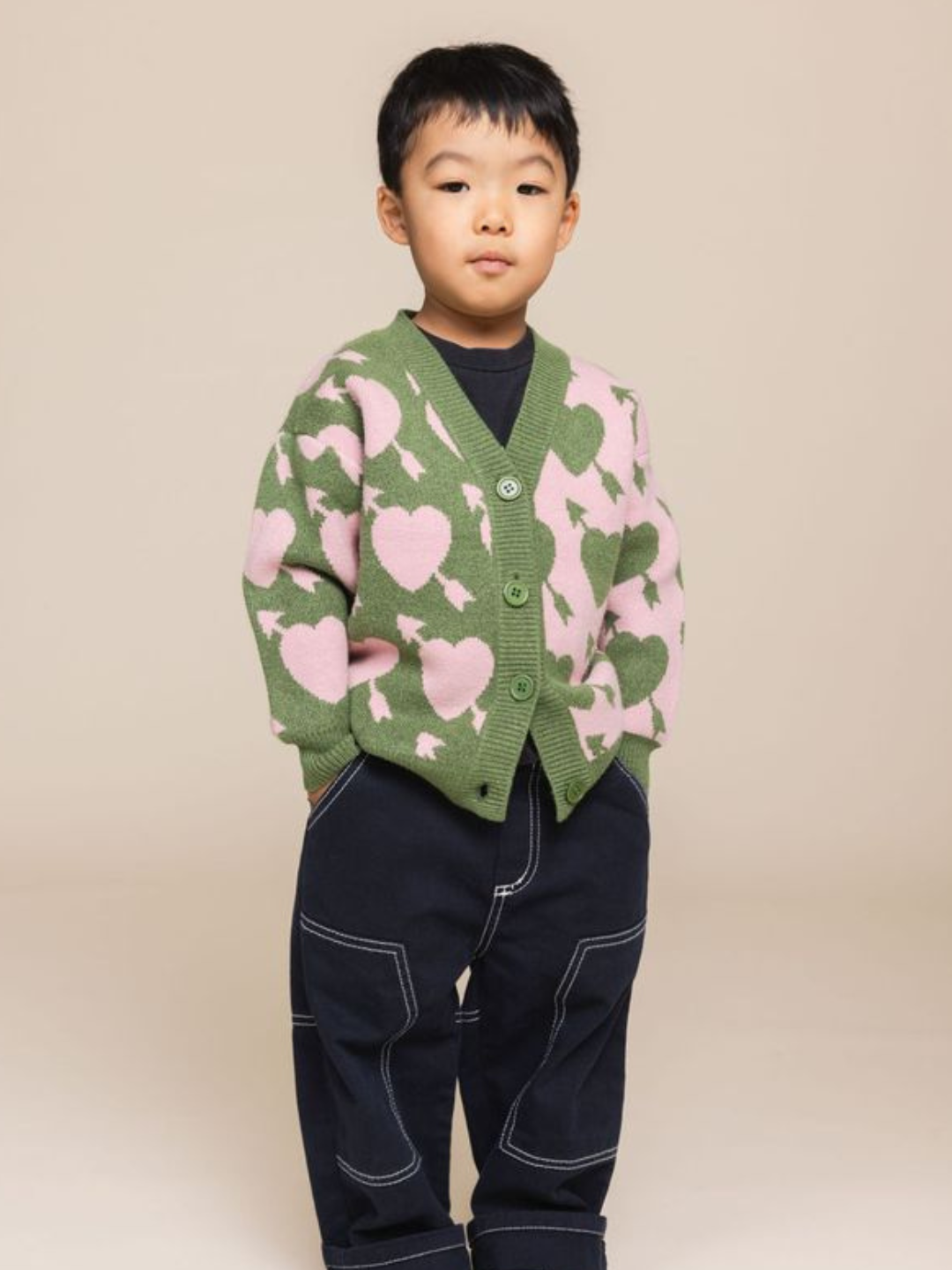 A child stands against a beige background, wearing the LOVESTRUCK CARDIGAN with its green and pink heart pattern, paired with dark jeans featuring white stitching. The child has a neutral expression and hands tucked in the pockets of the jeans. This charming photo captures every detail effortlessly.