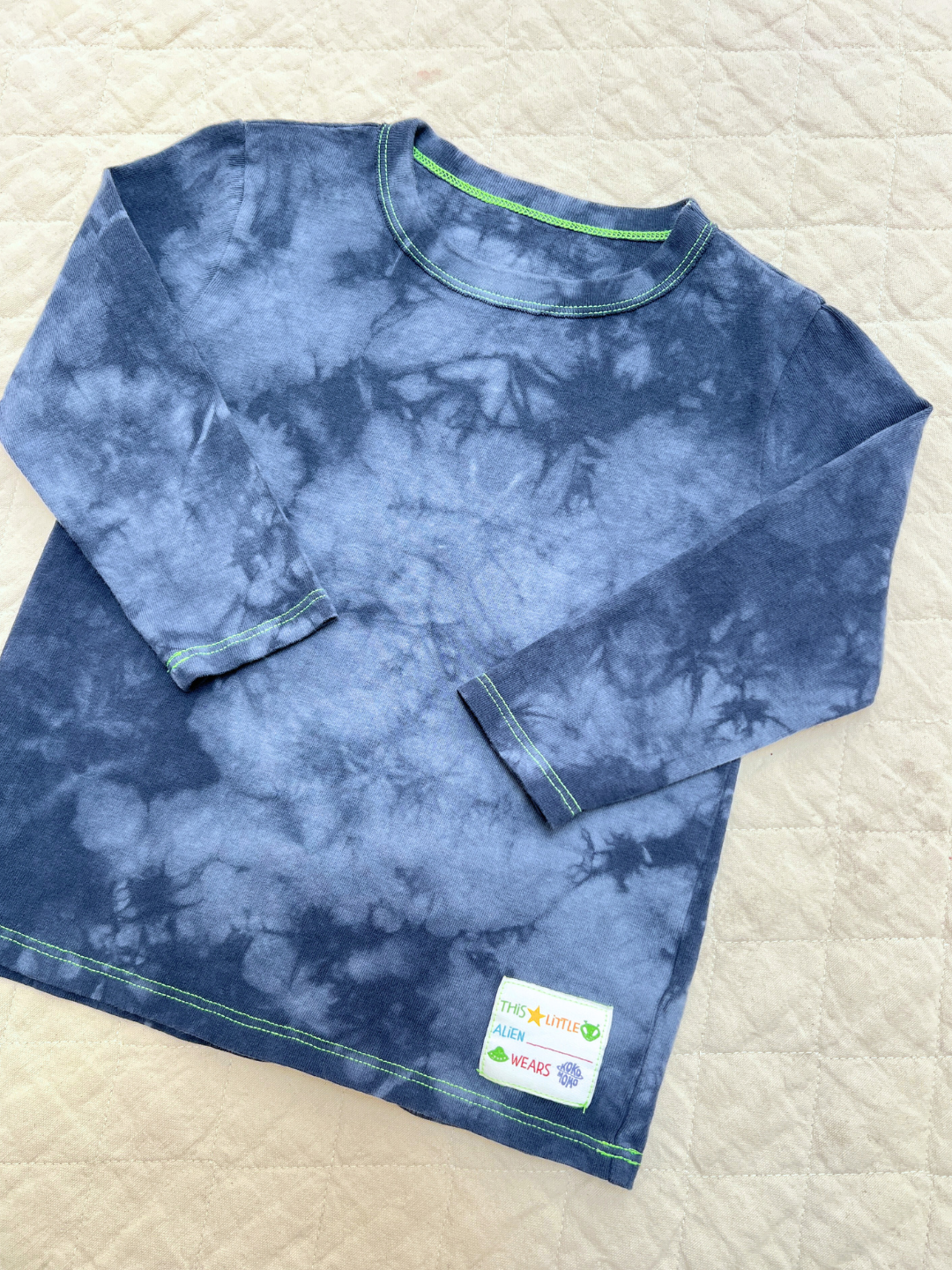 Pacific Blue | The COSMO TIE DYE LONG SLEEVE, featuring blue tie-dye with green stitching and a small label near the hem, is displayed flat on a quilted beige surface—channeling a bold Los Angeles-inspired look.