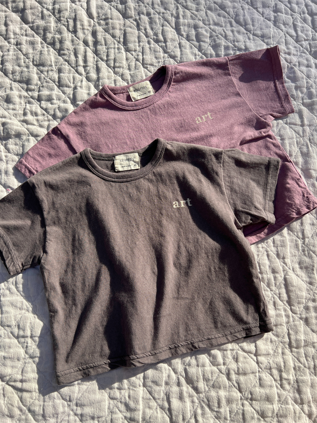 Charcoal | Two kids' Studio tees laid flat on a quilt in the sun