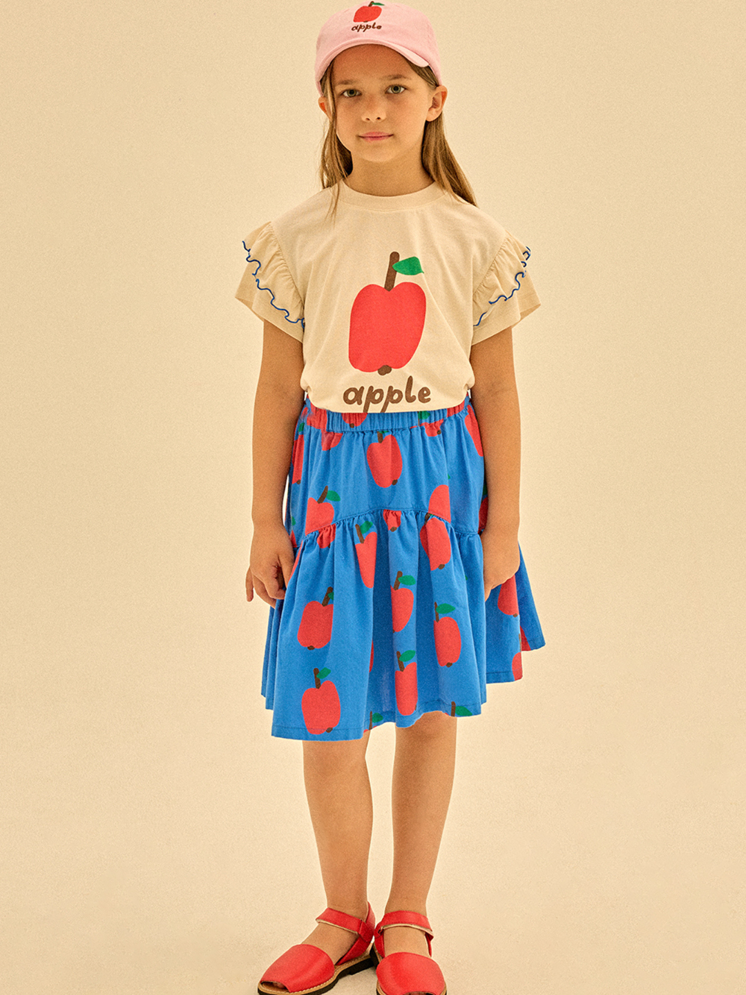 A girl in a beige shirt with apple design, an APPLE SKIRT (blue with red apples), red shoes, and a pink cap featuring an apple stands against a plain background.