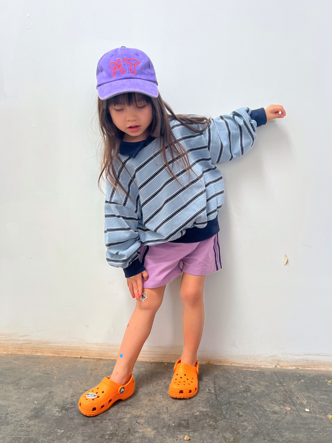 Purple | A young girl stands indoors against a white wall, wearing the MT DAD CAP (purple), a striped sweatshirt, cotton canvas purple shorts, and orange clogs. She bends one knee and looks slightly downward.