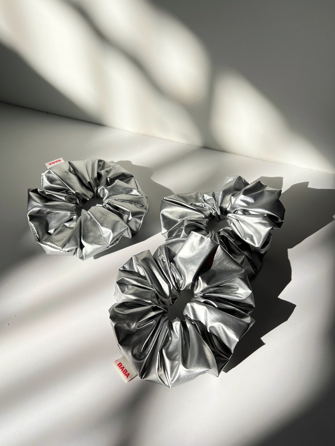 Three silver metallic scrunchies with red "Baba" logo tag on the side, lying on a white backdrop with shadows from a window..