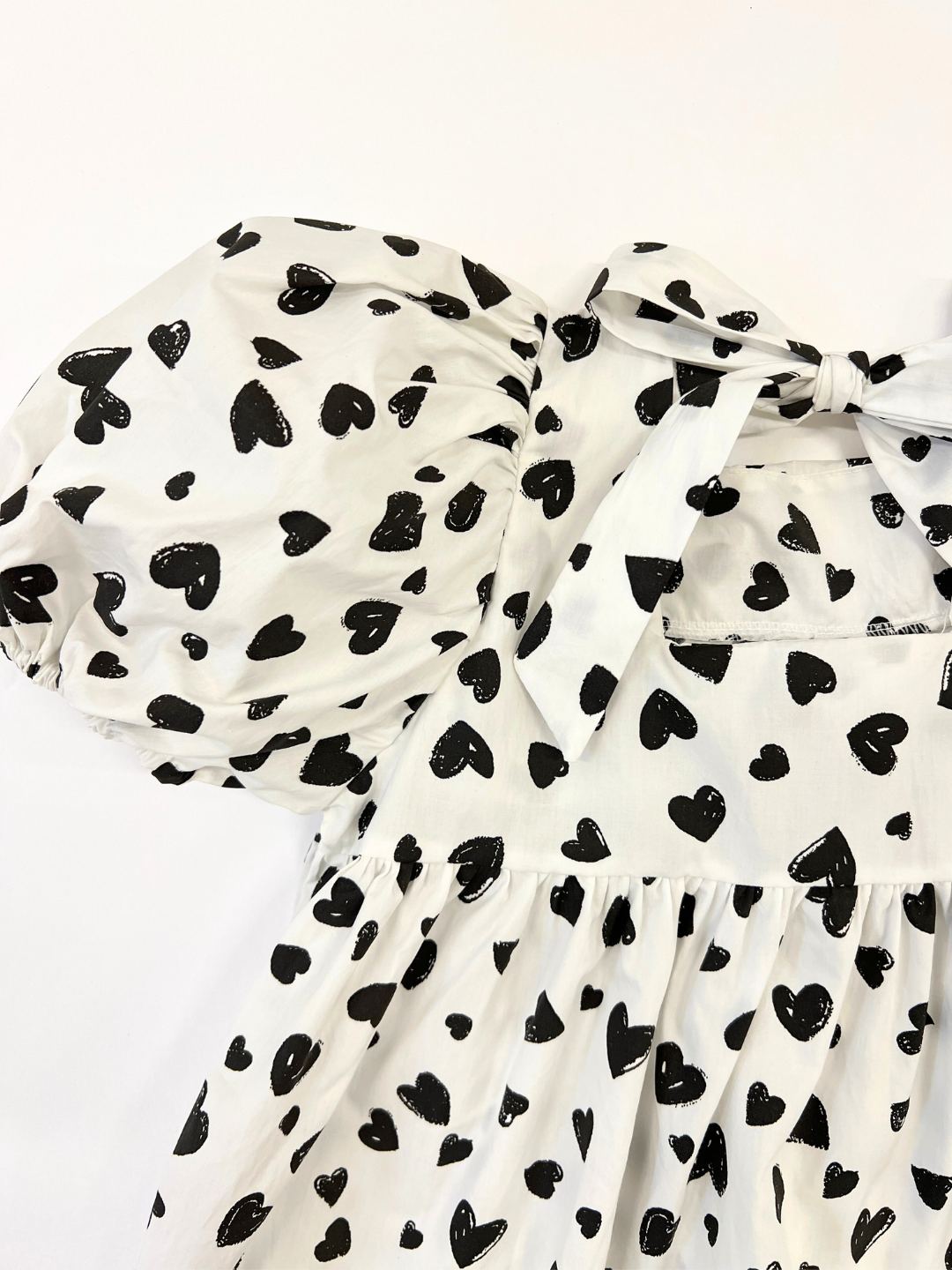 Close-up of the puff sleeve on a kids' dress in a pattern of black hearts on a white background, showing bow tied at neck, back view