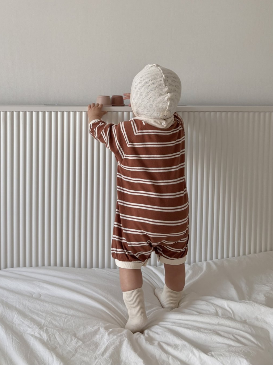 Terracotta | A baby stands on a bed facing a white headboard, wearing the SLOUCHY STRIPE ROMPER—a brown and white striped shortie made of soft cotton jersey from a Korean baby brand.
