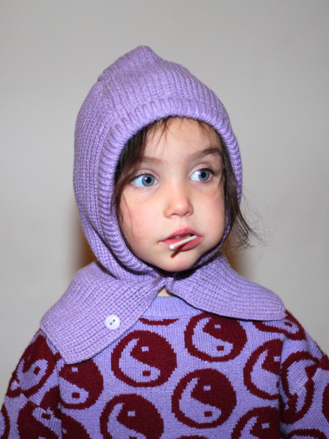 Purple | Child wearing the hooded hat in purple, with a purple sweater patterned with burgundy yin yangs. She is eating a lollipop and standing in front of a gray wall.