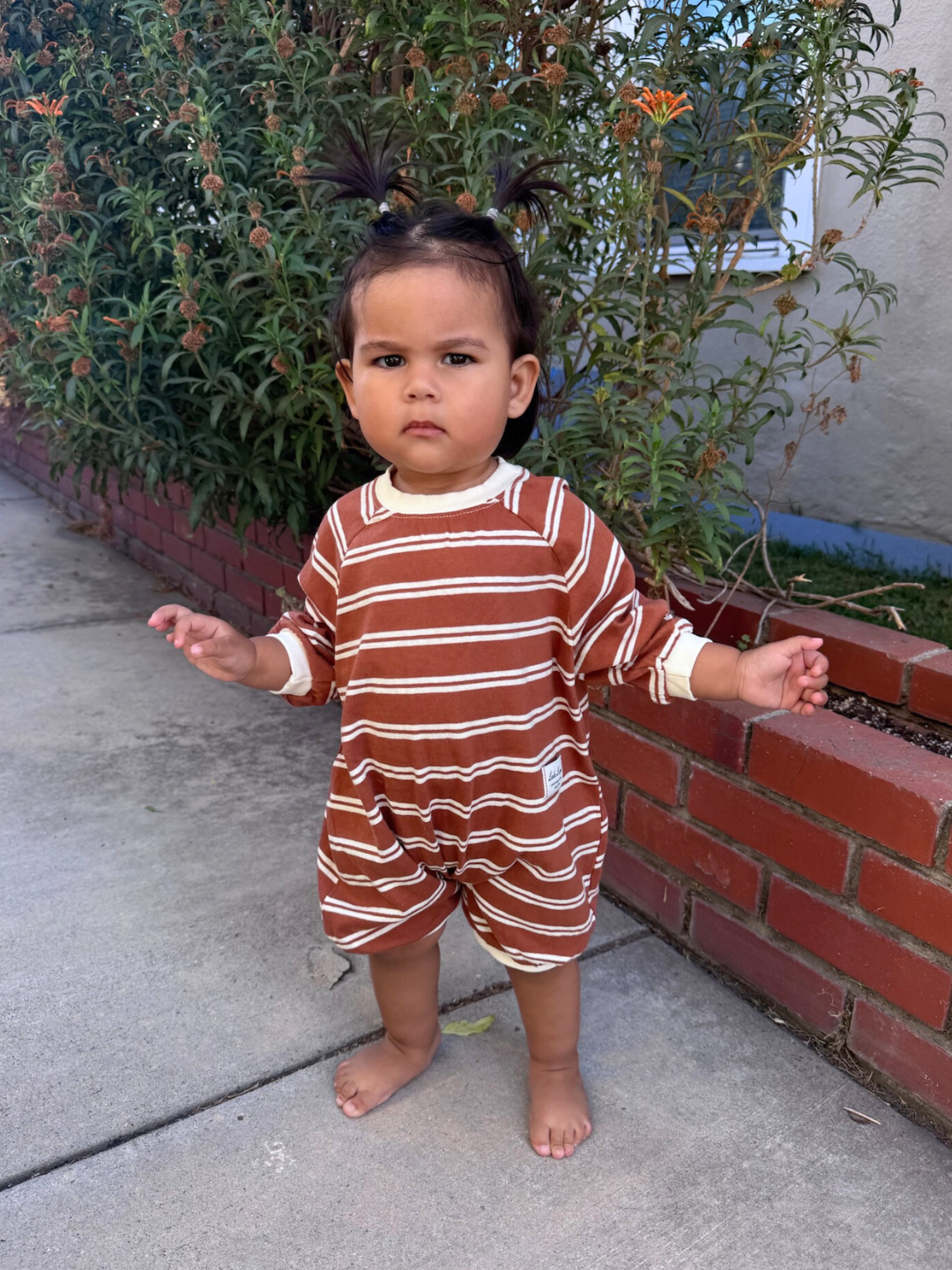 Child wearing a brown and white striped outfit standing on a sidewalk with plants in the background