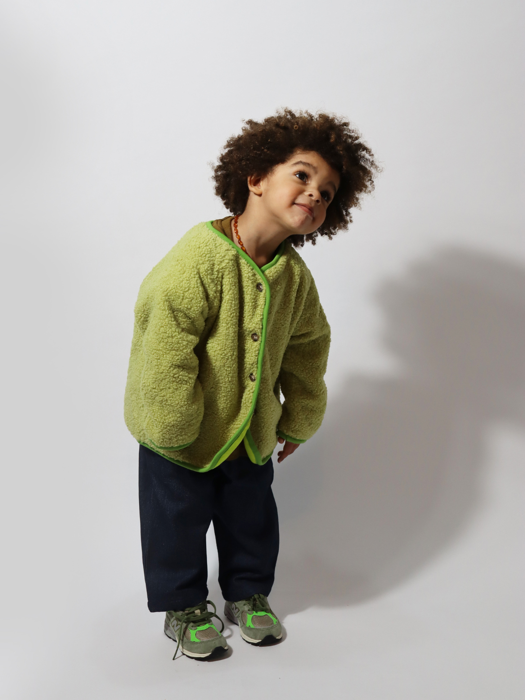 Matcha | A toddler wearing a light green collarless fleece jacket with four brown buttons, with dark blue pants and green sneakers, standing on a light grey background.