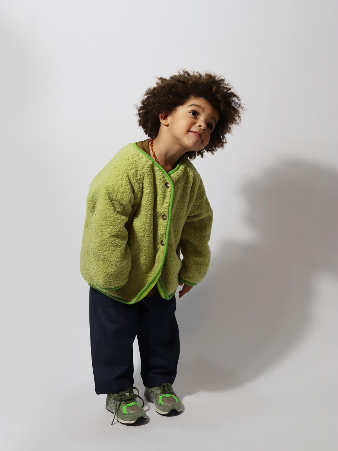 A toddler wearing a light green collarless fleece jacket with four brown buttons, with dark blue pants and green sneakers, standing on a light grey background.