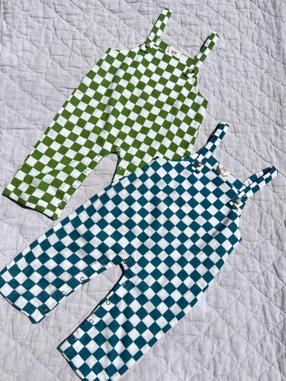 Two pairs of kids' overalls laid on a quilt; one in grass green and white check, the other aquamarine and white