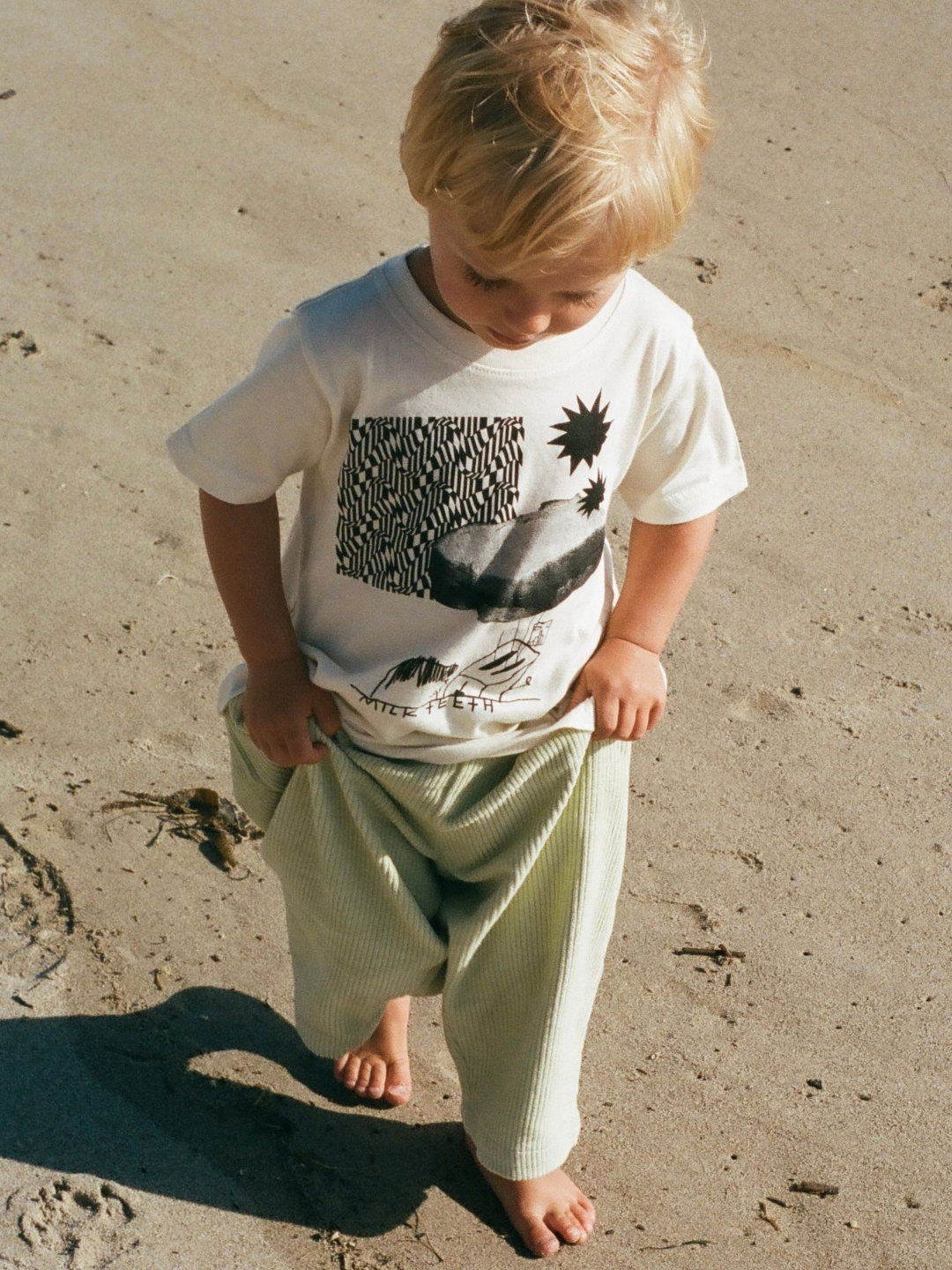 A child on a beach wearing a kids' tee shirt in cream, printed in black with patterns, stars, a PBJ sandwich and the words Milk Teeth