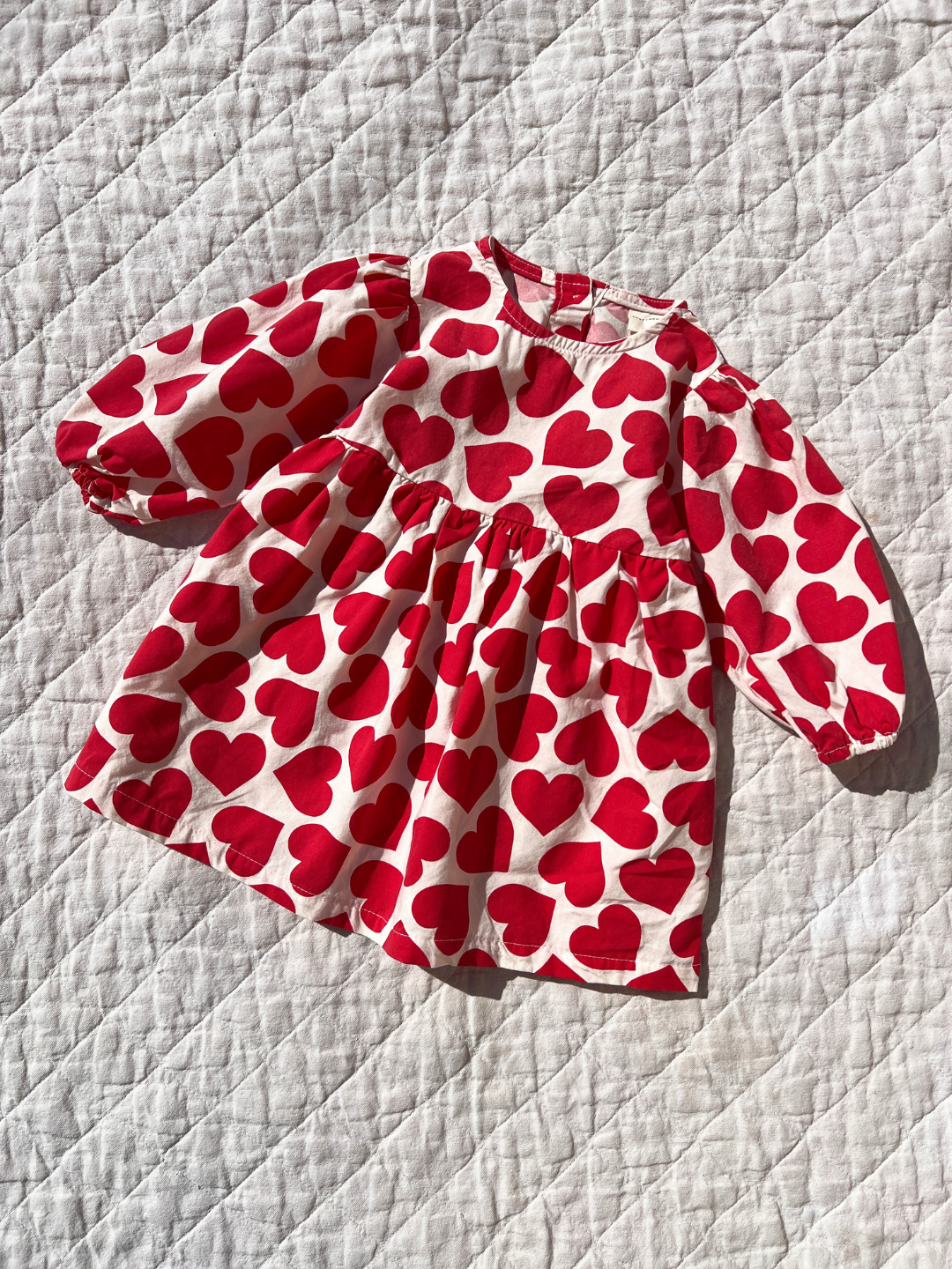 Kids dress in white with red hearts printed closely all over, with a round neck, high waist, long puffed sleeves and a loose, voluminous shape. Shown lying on an off-white quilt in sunlight.