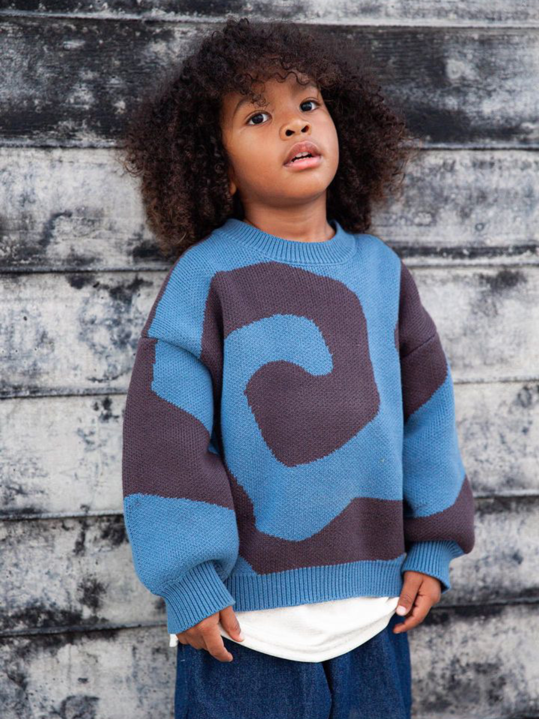 Slate | A child wearing a kids crewneck sweater in medium blue, with a large single charcoal grey swirl design that covers the entire sweater. He is standing against a grey wooden wall and also wears dark blue baggy jeans.