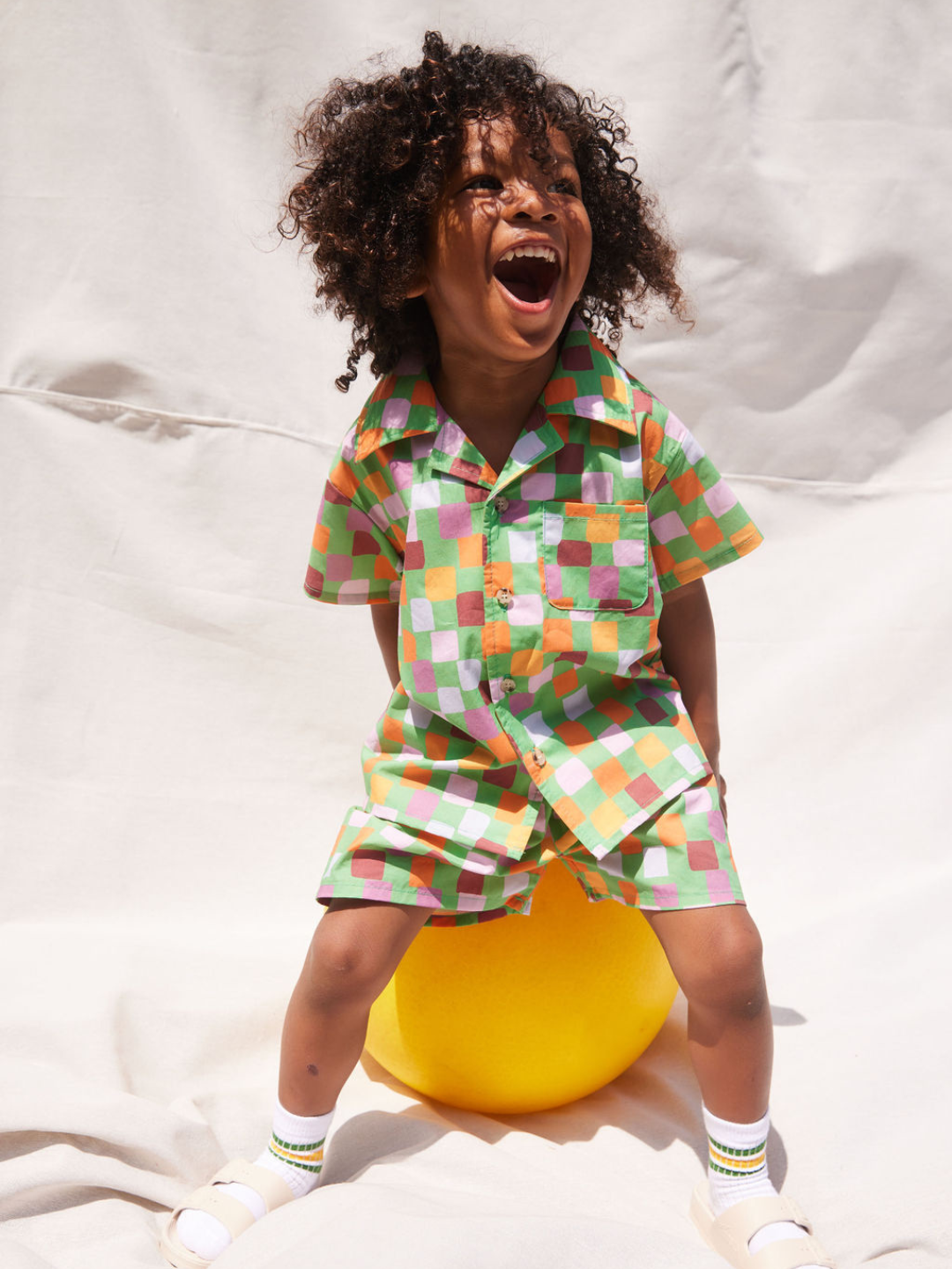 A child sitting on a ball, wearing a kids' shirt and shorts set in a pattern of purple, prink, gold and orange squares on a green background