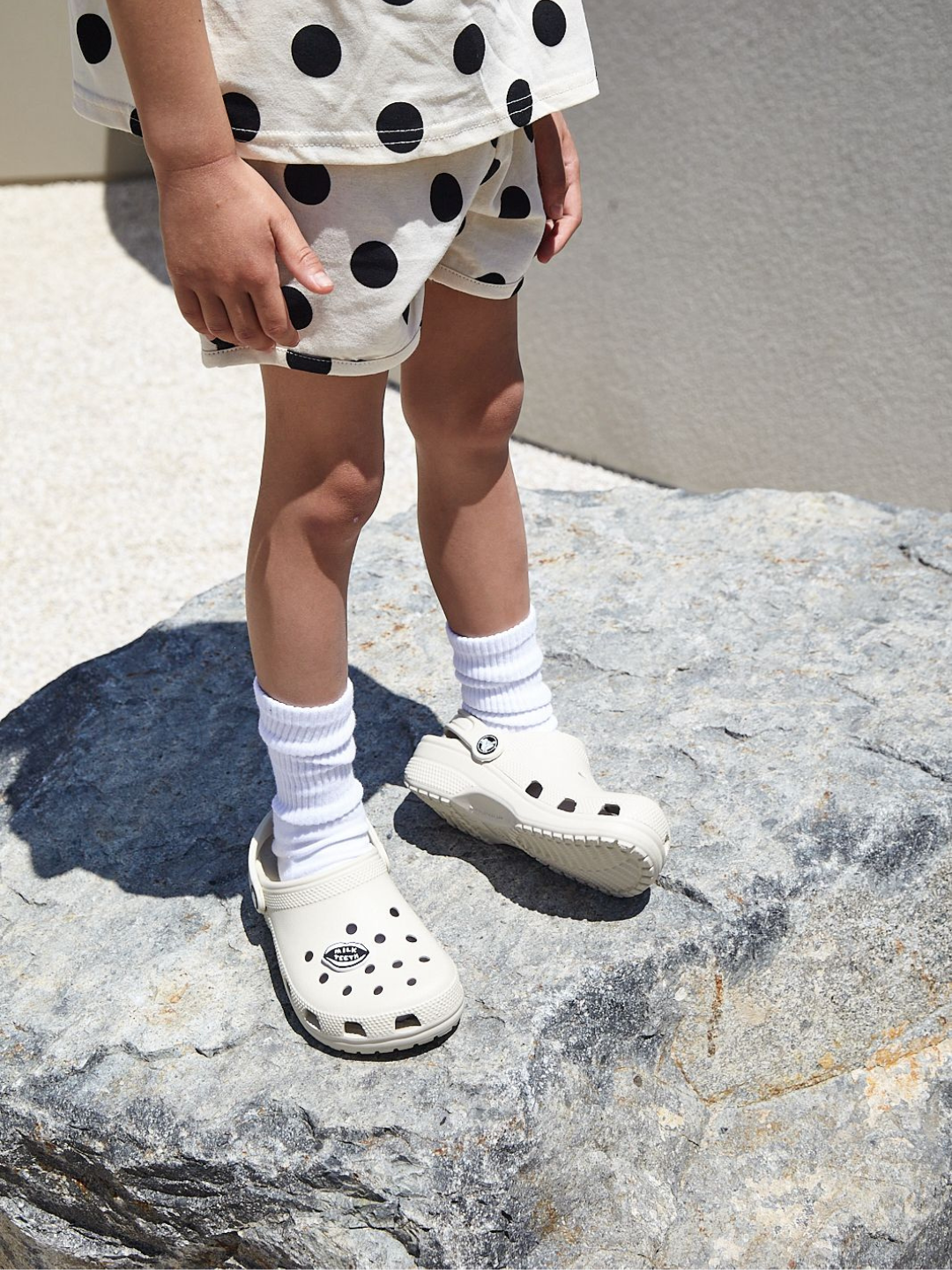  Close up of a child wearing a kids' tee shirt and shorts set in a pattern of black dots on an ecru background, with white Crocs and a Milk Teeth charm