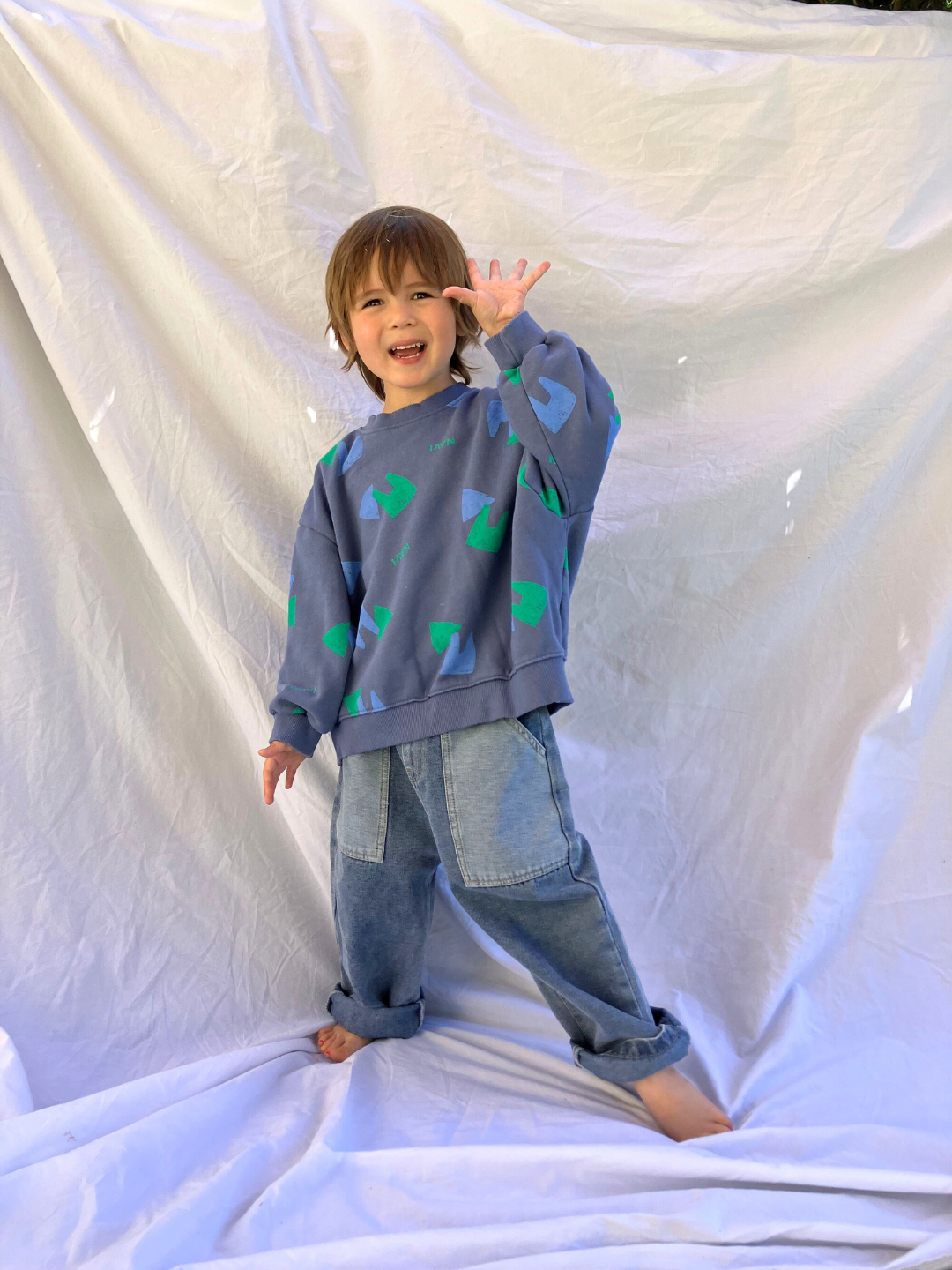 Vintage Blue | Child wearing the kids Duo Sweatshirt in faded blue printed with green and blue shapes and the word Navi. He is smiling and waving one hand, and wearing baggy blue jeans, and standing on a white sheet backdrop.
