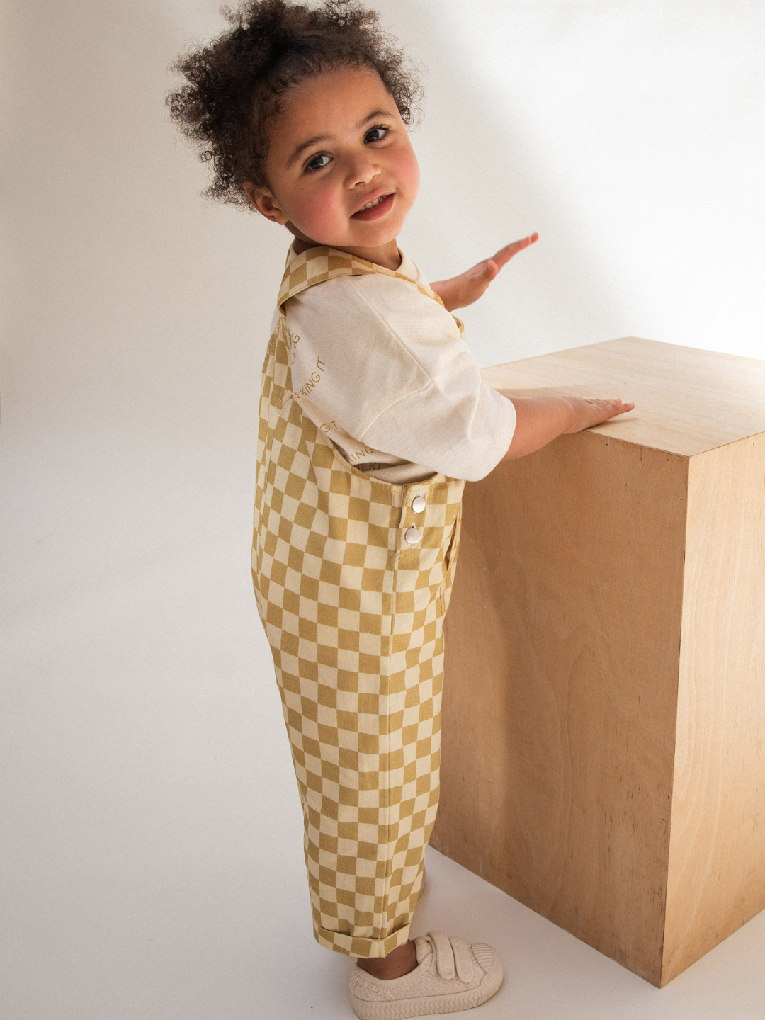 A young child with curly hair stands next to a wooden block, facing the camera. The child is wearing a white shirt under CHECK OCHRE DUNGAREE made of organic cotton, featuring a yellow and white checkerboard pattern and adjustable straps, paired with white shoes. The background is plain and well-lit, drawing attention to the child.