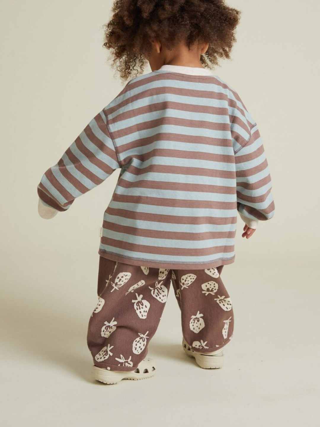A young child stands facing away, dressed in the RUE CHOC STRIPE LONG SLEEVE TEE and wide-legged brown pants with white insect prints.