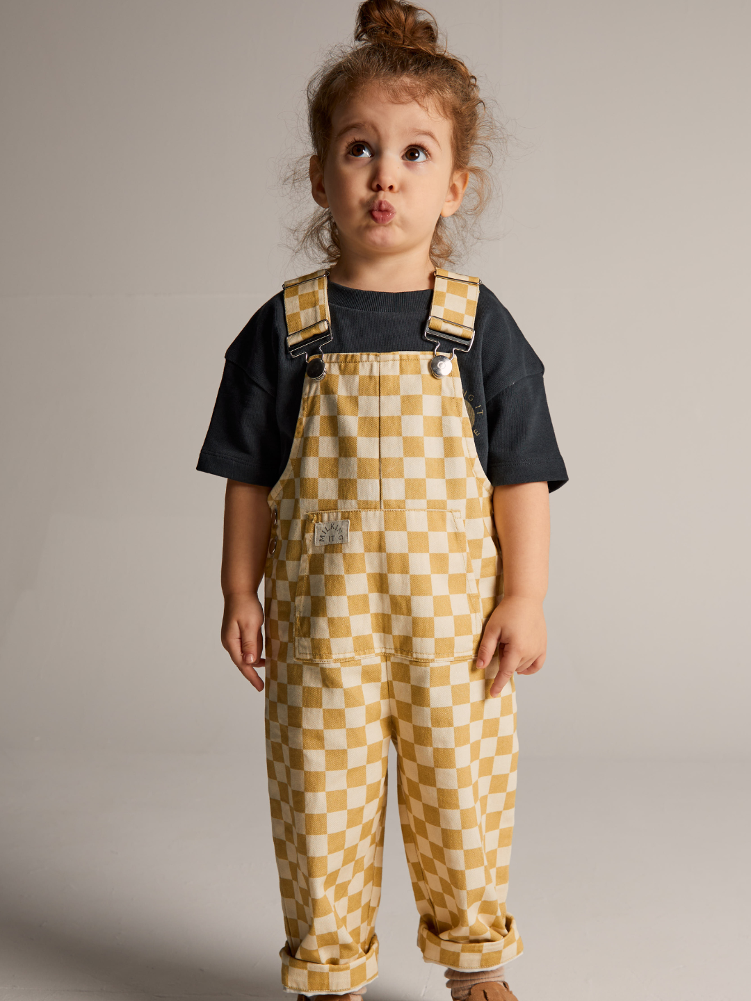 A young child with a bun hairstyle stands against a plain background, wearing Claude & Co's CHECK OCHRE DUNGAREE over a black T-shirt. The yellow and white checkerboard overalls made from 100% organic cotton complement the child's playful expression with puckered lips.