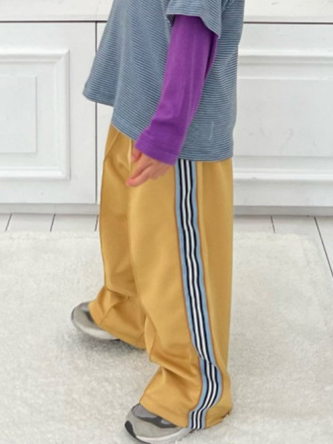Yellow | A child wearing MASKING TAPE PANTS—yellow with blue and white stripes—pairs them with a purple long-sleeve shirt under a striped tee and gray sneakers while standing on a light carpet.