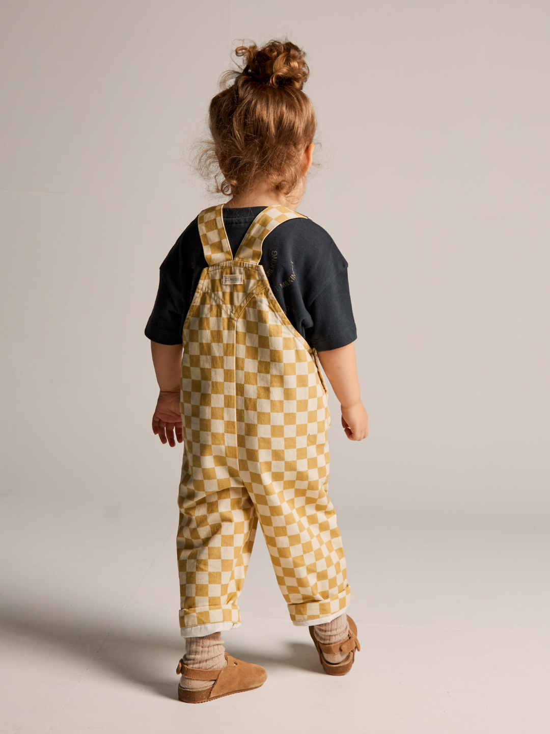 A toddler stands facing away from the camera, sporting a black shirt under Claude & Co's Check Ochre Dungaree, crafted from 100% organic cotton in a yellow and white checkerboard pattern. The child has light brown hair tied in a messy bun and is wearing light brown shoes. The plain, light-colored background enhances the focus on the child.