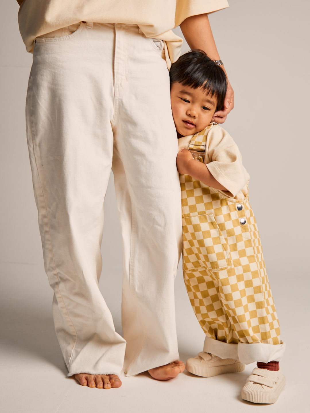 A small child wearing the Check Ochre Dungaree from Claude & Co and beige shoes stands closely next to an adult, dressed in light-colored pants and a light shirt. The child is leaning against the adult's leg, showing a sense of comfort and security.