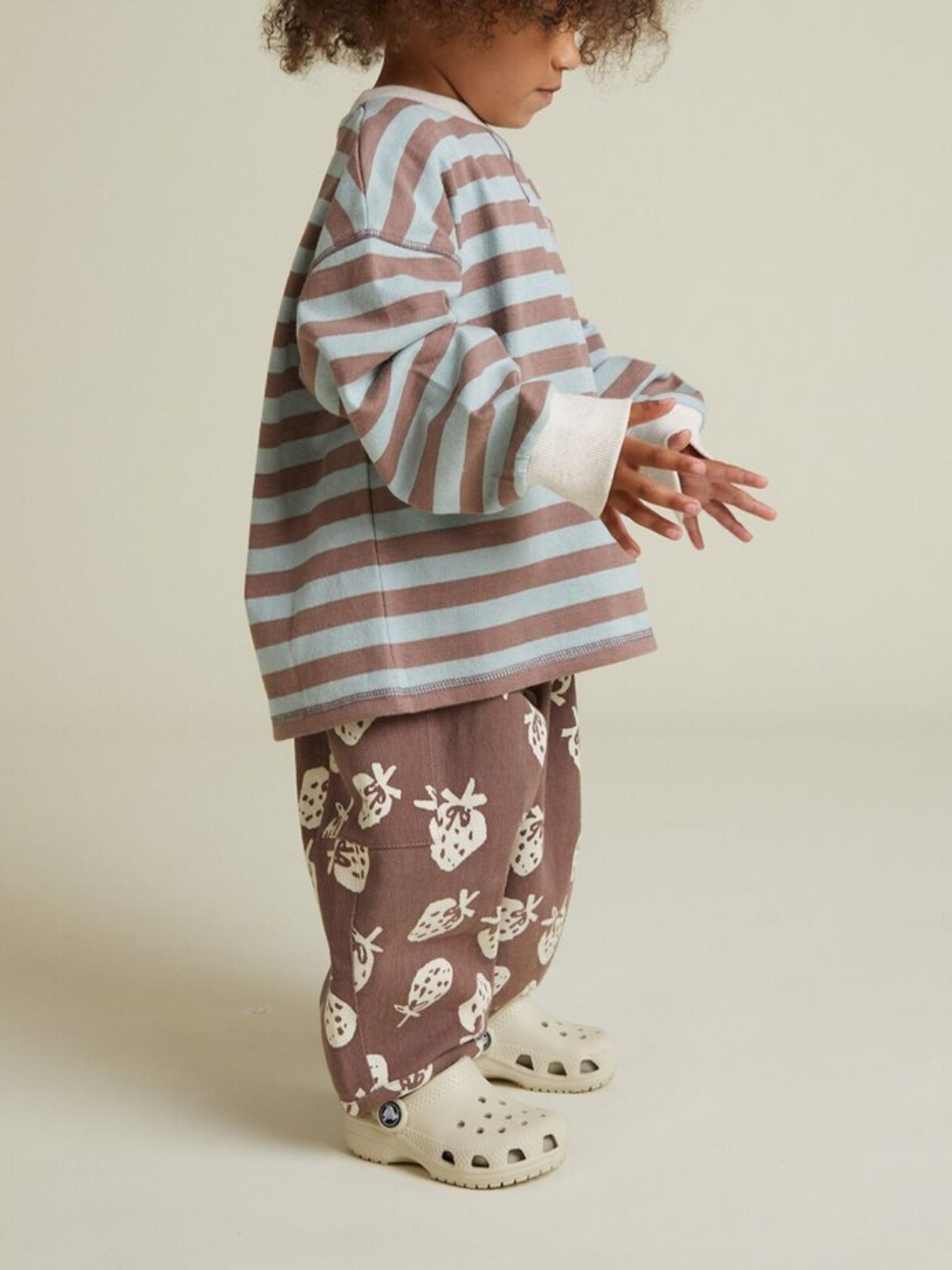 A young child stands against a plain background wearing the RUE CHOC STRIPE LONG SLEEVE TEE, brown patterned pants, and light rubber clogs.
