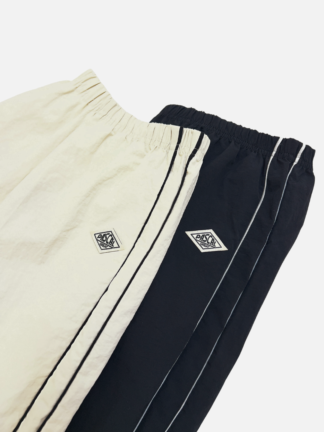 Set of two MINI WARMUP PANTS—one off-white, one black—each with an elastic waistband, sporty side stripes, and a diamond-shaped logo on the left thigh. Shown against a clean white background.