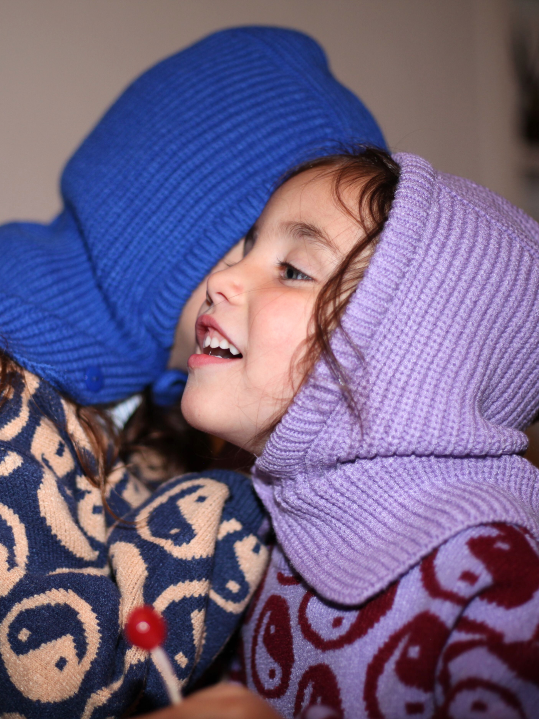 Two children wearing the hooded hats. One child wears a blue hat and a blue sweater and is whispering in the ear of a smiling child in a purple hat and sweater, who holds a lollipop.