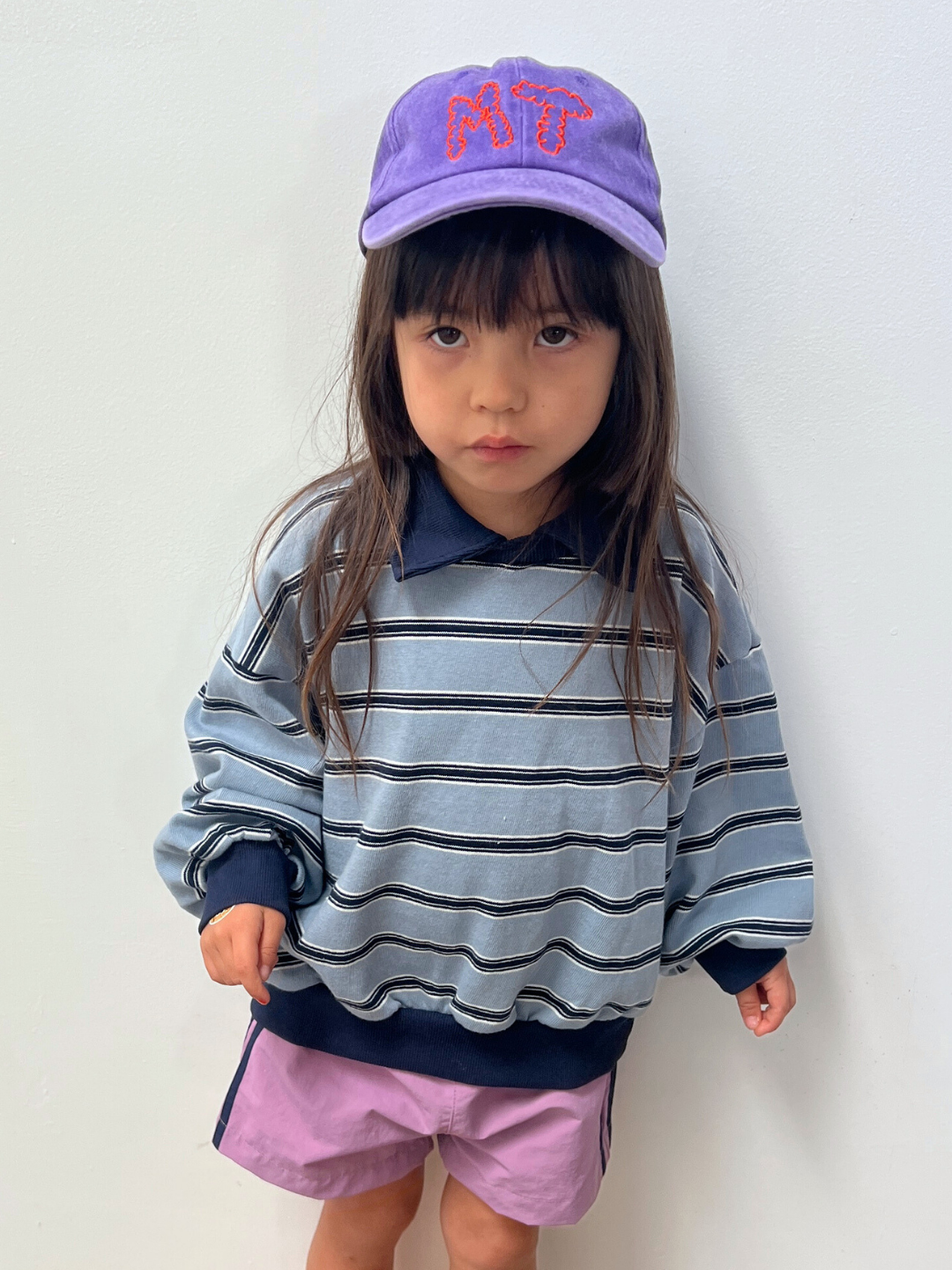 Purple | A young child with long brown hair wears a striped sweatshirt, purple shorts, and the MT DAD CAP—a cotton canvas baseball cap with red embroidered letters—standing against a plain white background.