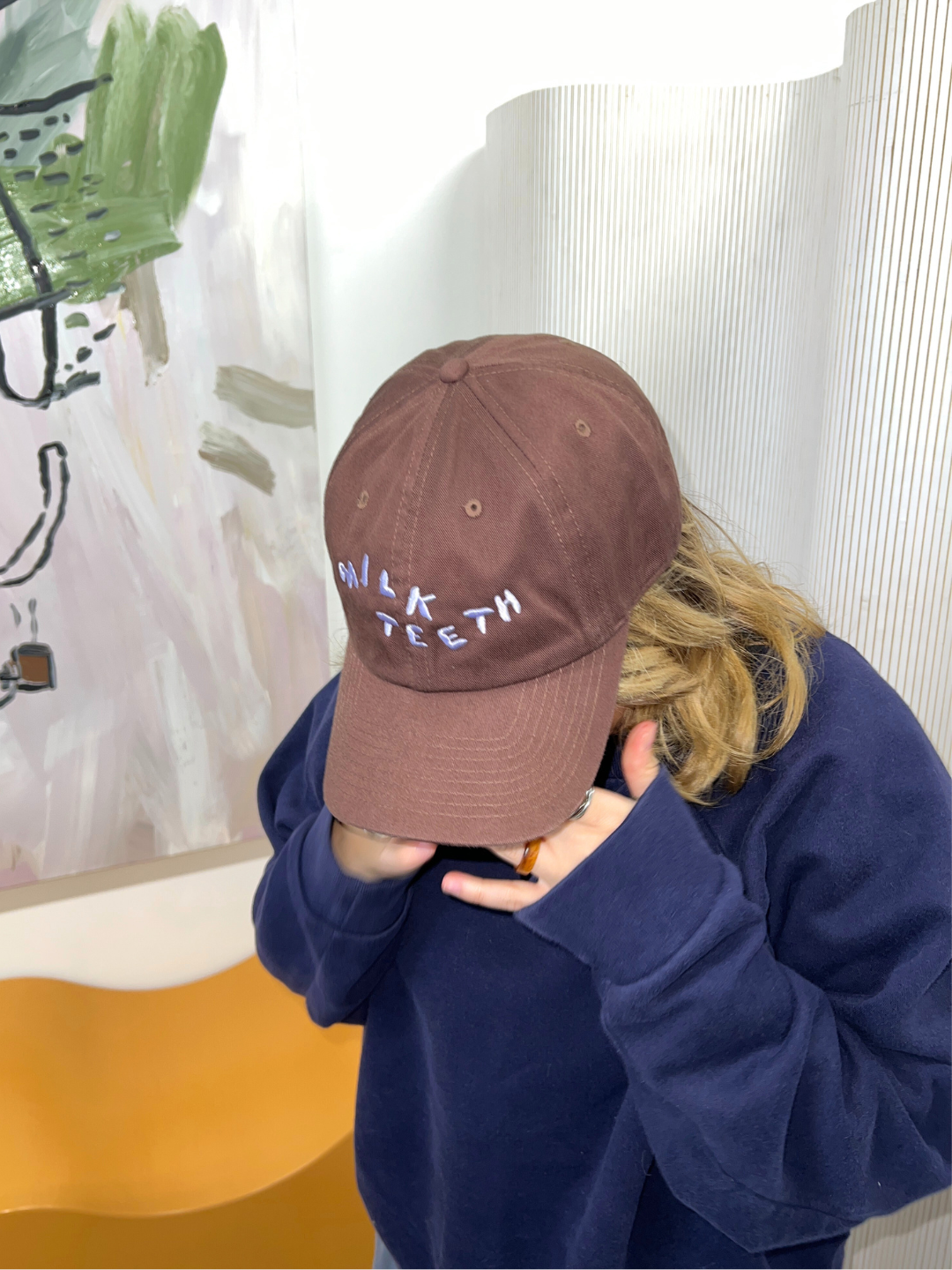 An adult wearing the Milk teeth adult baseball cap in brown with lavender logo embroidery. They wear a navy sweatshirt in a white room. Behind them is an orange bench and an abstract painting.