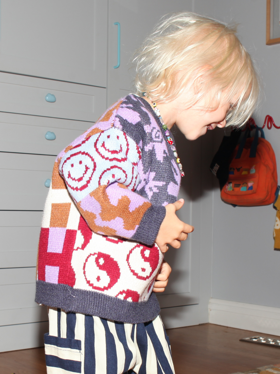 A young child with blond hair, wearing a colorful sweater and striped pants, stands indoors by a dresser featuring the handmade GAIA NECKLACE with strawberry and mushroom beads, next to a wall with hanging bags.