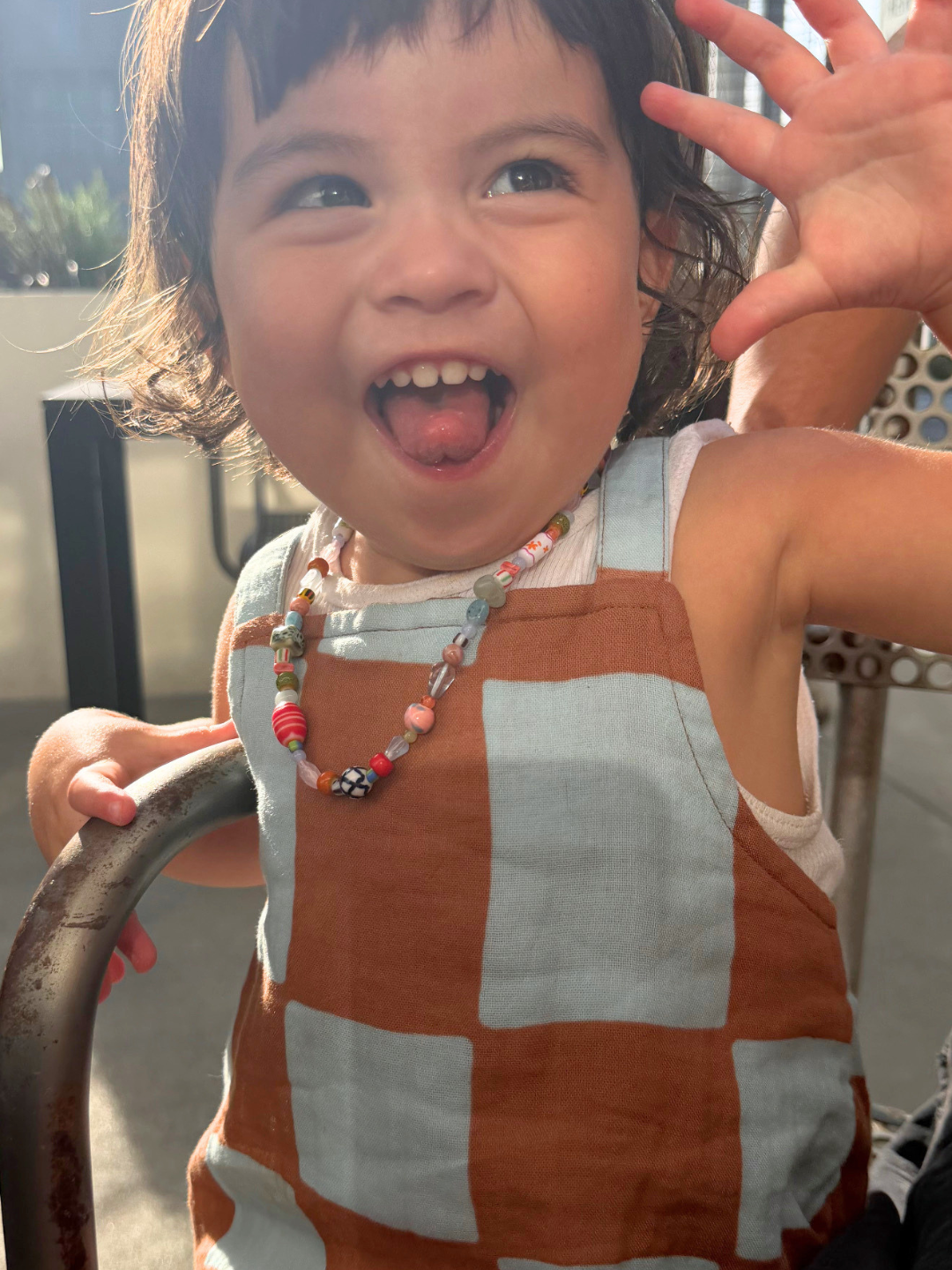Blue Checkers | Child wearing a blue and brown checkered romper with a necklace, waving outdoors.